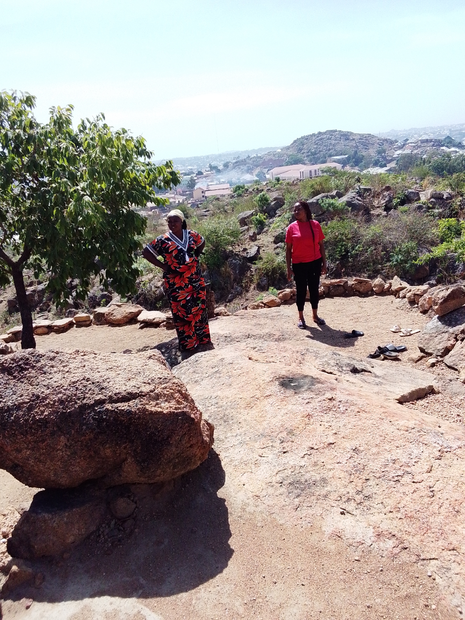 Two women stand on a rocky plateau overlooking Gura Top, Nigeria, under a bright, cloudless sky. One woman wears a colorful, floral-patterned dress and a headwrap. The other woman is dressed in a red t-shirt and black leggings. Several pairs of sandals lie on the ground near the second woman. A small tree grows to the left of the first woman, and a large boulder sits in the foreground. In the distance, the landscape is dotted with buildings and rolling hills.