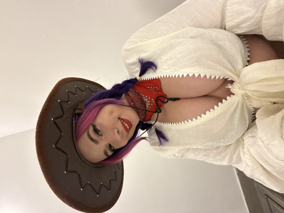 A woman with pink and purple hair, wearing a cowboy hat, faces the camera. She has red lipstick and dark eyeliner. A red bandana covers her neck. She is wearing a white garment with a notched edge and a tie, revealing cleavage. The picture is taken from a low angle, looking up at her. The background is a plain white wall.