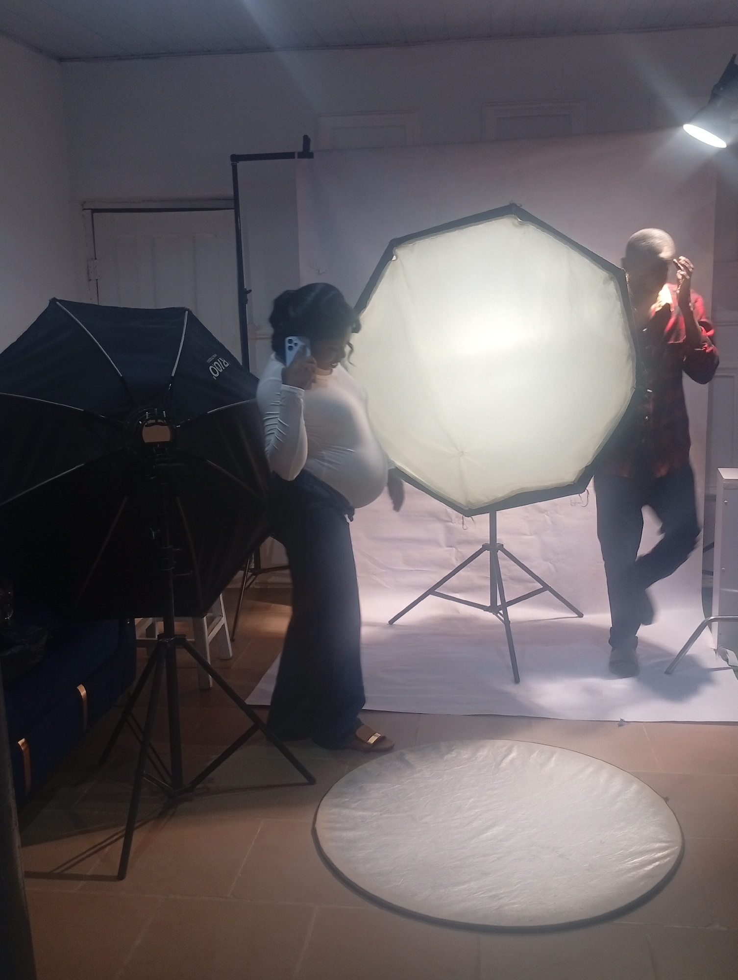 A pregnant woman is using a smartphone in a photography studio. She is wearing a light-colored long-sleeved shirt and dark pants. In the studio, there is a large black umbrella light with a tripod stand to her left and a large octagonal softbox on a tripod stand to her right. A man in a red plaid shirt and dark pants is standing partially obscured by the softbox, adjusting his hair or glasses. A white backdrop is behind them, with a light shining on it from above. On the floor, there is a circular reflector. The studio appears to be in a room with a tiled floor and a partially visible blue couch on the left side of the frame. The location is Jos, Nigeria.