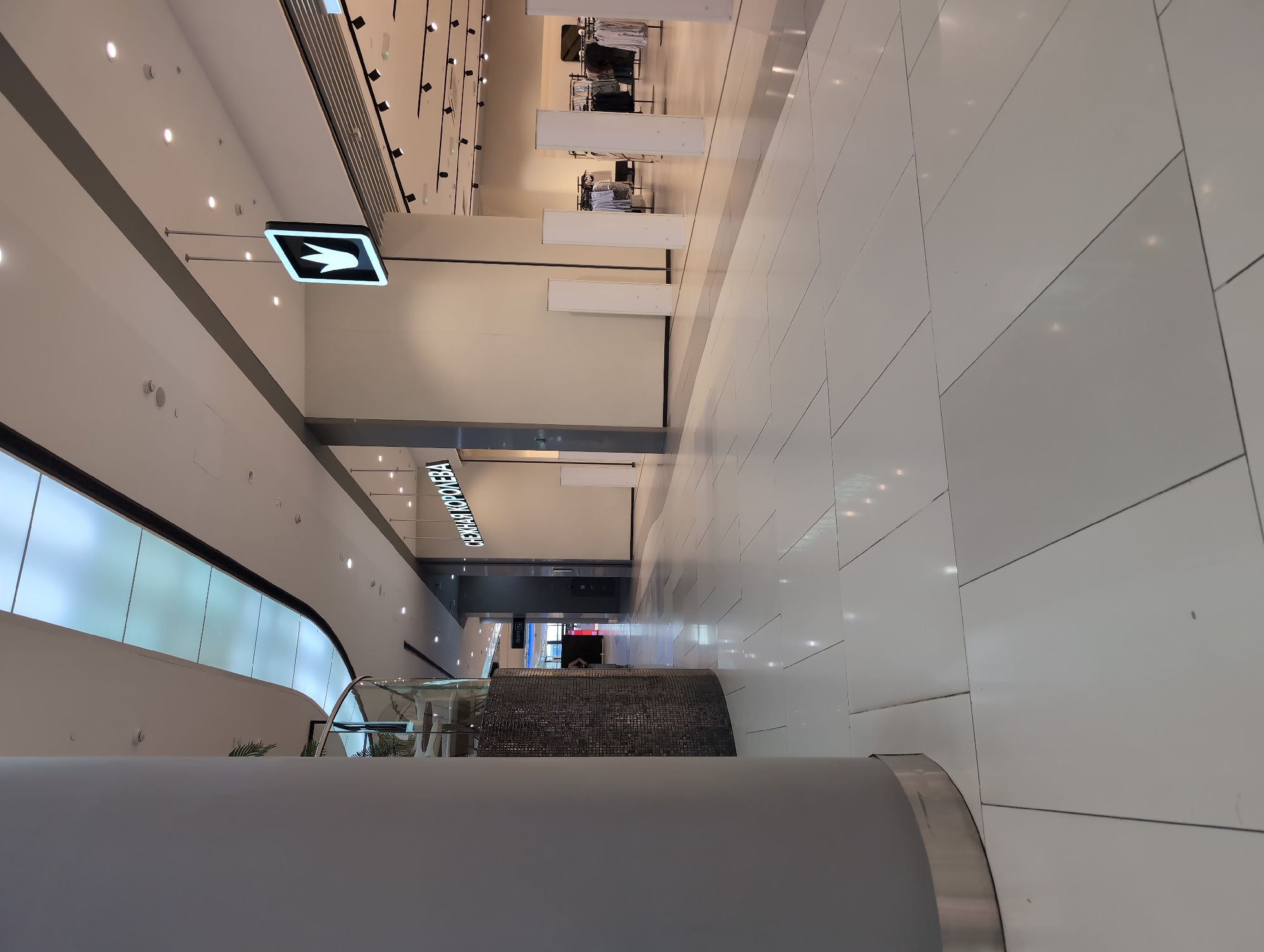 This media file captures an interior view of a modern shopping mall, likely located in Krasnodar, Russia. The scene depicts a wide, brightly lit corridor with a polished, light-colored tiled floor that reflects the overhead lighting. The architecture is contemporary, featuring a high ceiling with numerous spotlights and a sleek, curved facade on the left side, possibly housing shops or displaying advertisements behind a translucent material.
Dominating the foreground on the left is a large, grey, cylindrical structure, possibly a pillar or part of an escalator. In the middle ground, a glass-sided escalator with chrome railings is visible, leading upwards. A decorative element with a mosaic-like texture is situated near the escalator.
Further down the corridor, signage is present. A prominent black and white directional sign with a white arrow pointing to the right is mounted on the ceiling. On the right wall, near the end of the visible corridor, there is a brightly lit sign with text that appears to be in Cyrillic script. Partial text reads "СНЕЖНАЯ КОРОЛЕВА" which translates to "Snow Queen", a known Russian retail chain. Other shop entrances or displays are visible in the distance, some with clothing racks.
The lighting suggests it is daytime, but the interior setting makes it irrelevant to natural light conditions. There are no people prominently featured in the image, giving the scene a sense of quietude and emptiness. The overall impression is one of a clean, spacious, and modern commercial environment.
