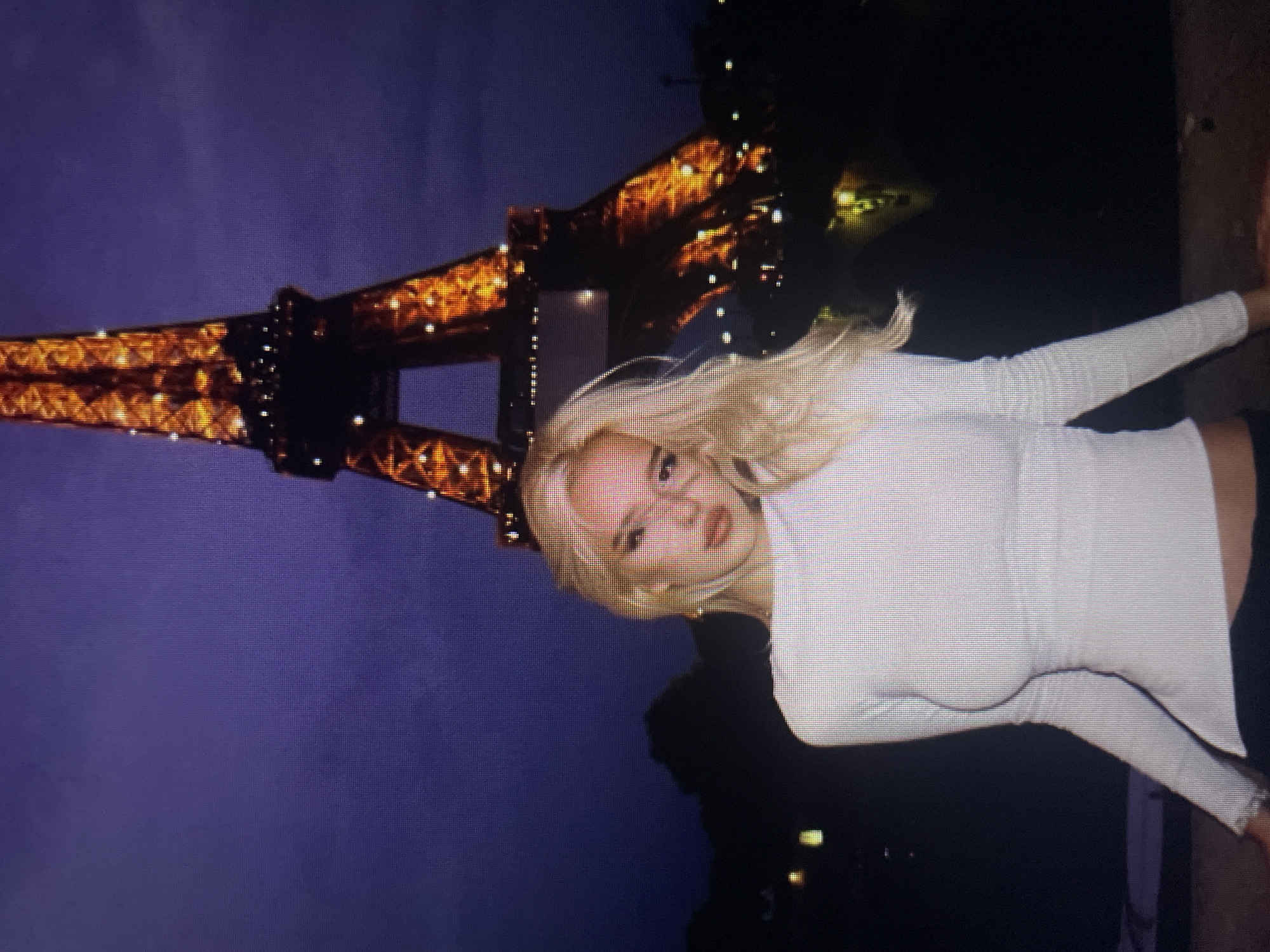 A young woman with blonde hair and light skin is posing in front of the Eiffel Tower at night. She is wearing a white long-sleeved shirt and black pants. The Eiffel Tower is illuminated with golden lights, and the sky is dark blue. The image appears to be taken in Paris, France, not Kołobrzeg, Poland.