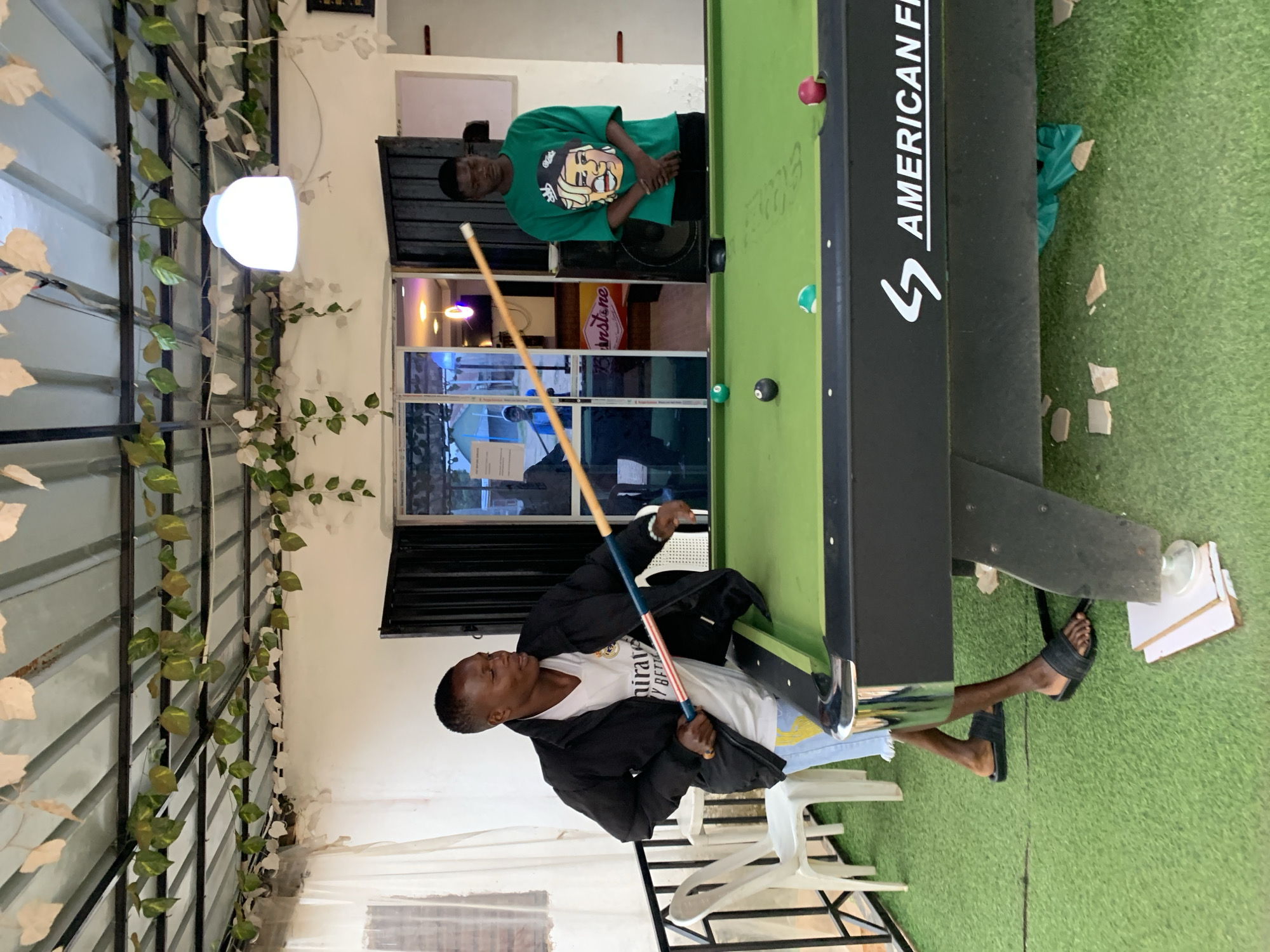 A man is playing pool at a table indoors. Another man in a green shirt stands by the side of the pool table, observing. The pool table is green with an American Flag logo on the side. The man playing pool wears a black jacket, a white T-shirt and jeans. He has a pool cue in his hand, and he is aiming at a ball on the pool table. The pool table is on a green surface. White chairs are present in the foreground. The location is suspected to be Jos, Nigeria.
