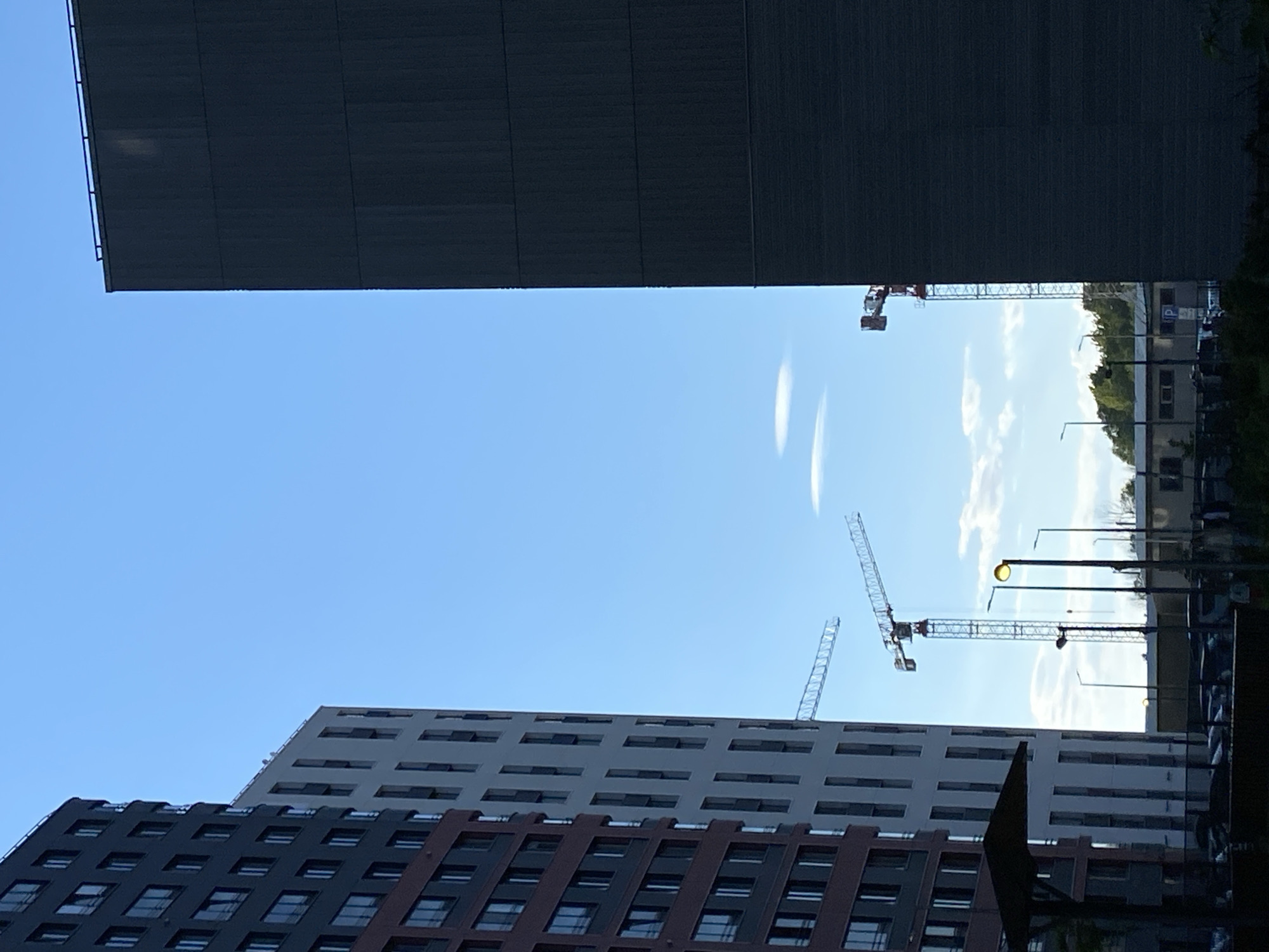 The image depicts a scene in Saint Petersburg, Russia, with a view between two buildings towards the sky. The upper building is a large, dark, rectangular structure. The lower building features a facade with alternating dark and light horizontal sections, punctuated by windows. Construction cranes are visible in the space between the buildings, indicating ongoing development. Street lights, foliage, and parked vehicles are also visible. The sky is blue with some wispy clouds.