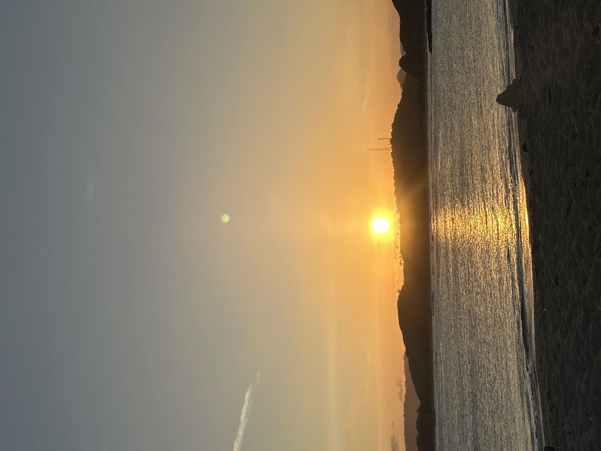 The image depicts a serene sunset over the ocean in Kuta, Indonesia. The sun, a brilliant orb of orange and yellow, is low on the horizon, casting a shimmering golden path across the water. Silhouetted against the vibrant sky are rolling hills and mountains, dotted with trees and what appear to be communication towers. The foreground is a sandy beach, with the dark sand contrasting with the gentle waves lapping at the shore. A single piece of driftwood rests on the sand, adding a natural element to the scene. The sky transitions from a pale blue at the top to a warm gradient of orange and yellow as it nears the horizon, with a few wispy clouds adding texture. Small boats are visible in the distance on the water. The overall impression is one of peaceful tranquility and the natural beauty of a coastal sunset.