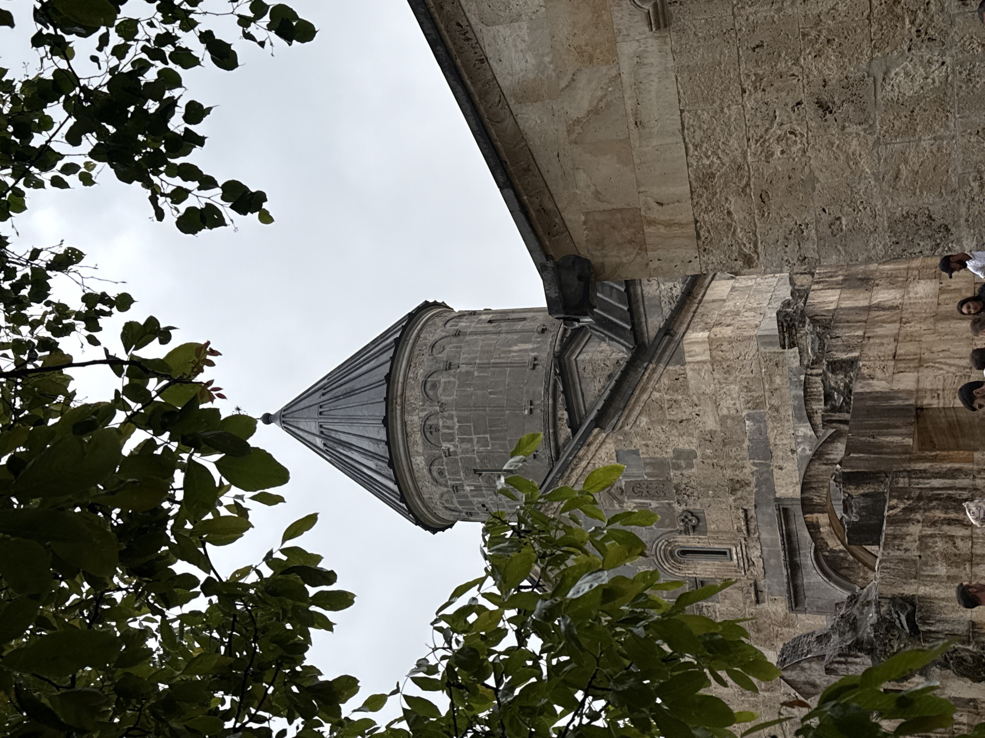 An exterior, medium shot captures a stone church structure with a conical roof visible through the branches of a tree. The church is constructed of rough-hewn stone blocks in shades of beige and gray. The main section of the building has a domed roof topped with a spire, and intricate stone carvings adorn its facade. To the right of the main structure, a wall of lighter-colored stone extends upwards, with a dark gutter running along its top edge. In the foreground, the lower part of the church features arched entrances and a section of ruined stonework, partially obscured by foliage. Several individuals are present in the lower portion of the frame, observing the structure. The sky above is overcast and gray.