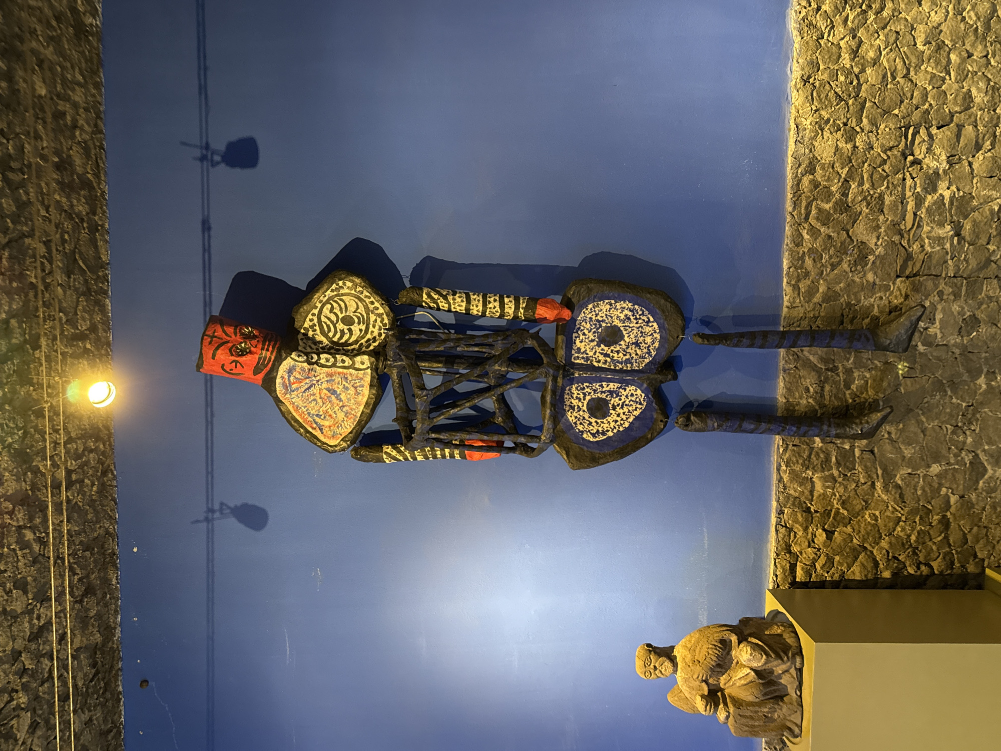 The image shows a wall in Mexico City, Mexico. The left side of the wall is a blue painted surface, while the right side is a rough stone wall. A decorative metal sculpture of a human figure is mounted on the blue wall. The figure has a red head, and its body is decorated with black, white, yellow, and blue patterns. The figure's legs extend onto the stone wall. Below the sculpture, there is a small, carved stone figure on a plinth. A light fixture is visible on the left side of the image, casting a shadow on the wall. Above the figure's head is a shadow cast by a hanging object.