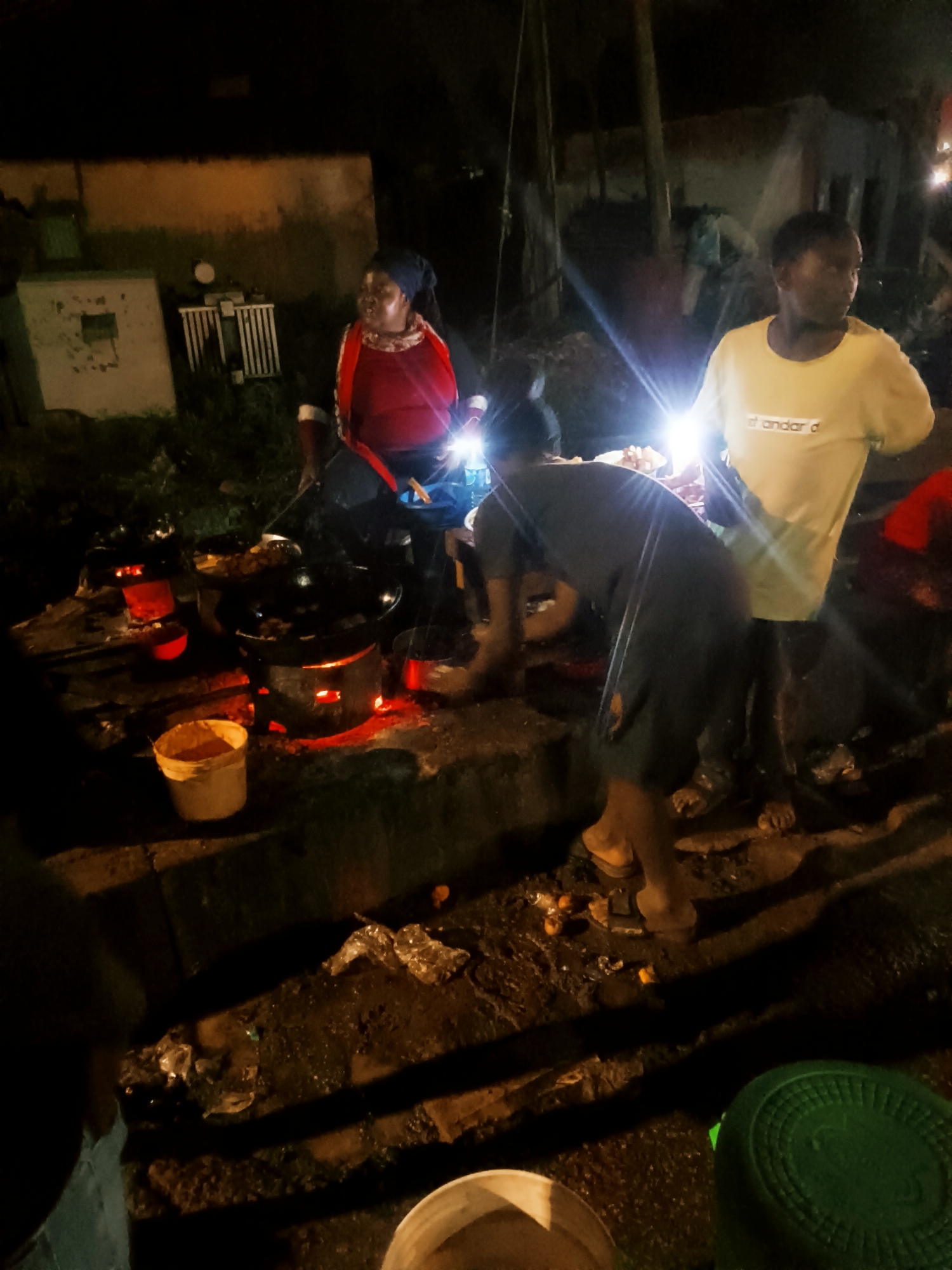 At night in Bukuru, Nigeria, three people are engaged in food preparation outdoors. A woman in a red shirt and head covering stands by several black pots sitting on red-glowing stoves. The stoves are placed on a surface built of stone or concrete. Another person wearing dark clothing bends toward the pots, and a third person in a yellow t-shirt is standing to the right. There is a bright light pointing toward the scene. To the left, there is a white building. The foreground shows the ground is dirty with scattered debris, and there are several plastic buckets visible.