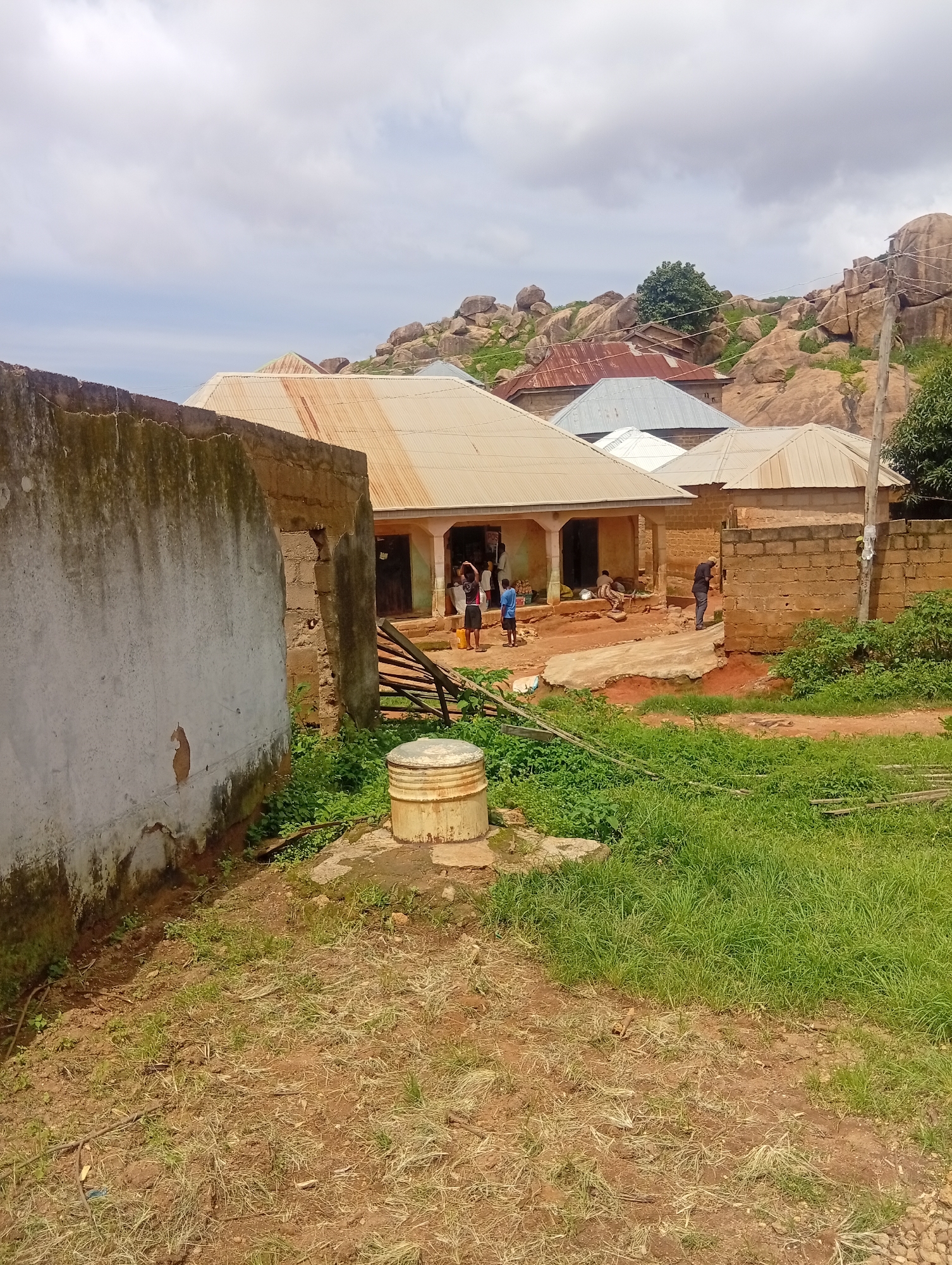 The image depicts a rural scene in Jos, Nigeria, characterized by a collection of simple, weathered buildings with corrugated metal roofs. In the foreground, a rough, unpaved yard area is visible, with patches of dry, sparse grass and scattered debris. To the left, a large, unfinished concrete wall, stained and cracked, dominates the frame.
Behind this wall, a low-slung building with a rusted metal roof extends towards the center of the image. Several people are gathered around the entrance of this building, including children playing and adults engaged in activities that suggest daily life, possibly commerce or domestic tasks. Further back, more buildings with similar roofing can be seen, some constructed with brick and others with what appears to be adobe or mud.
The environment is rugged, with a prominent backdrop of large, weathered rock formations and sparse green vegetation. The sky above is overcast with clouds, suggesting a possibly humid or rainy climate. A visible utility pole with wires is present in the middle ground, indicating some level of infrastructure in the area. The overall impression is one of a modest, functional settlement integrated into a natural, rocky landscape.