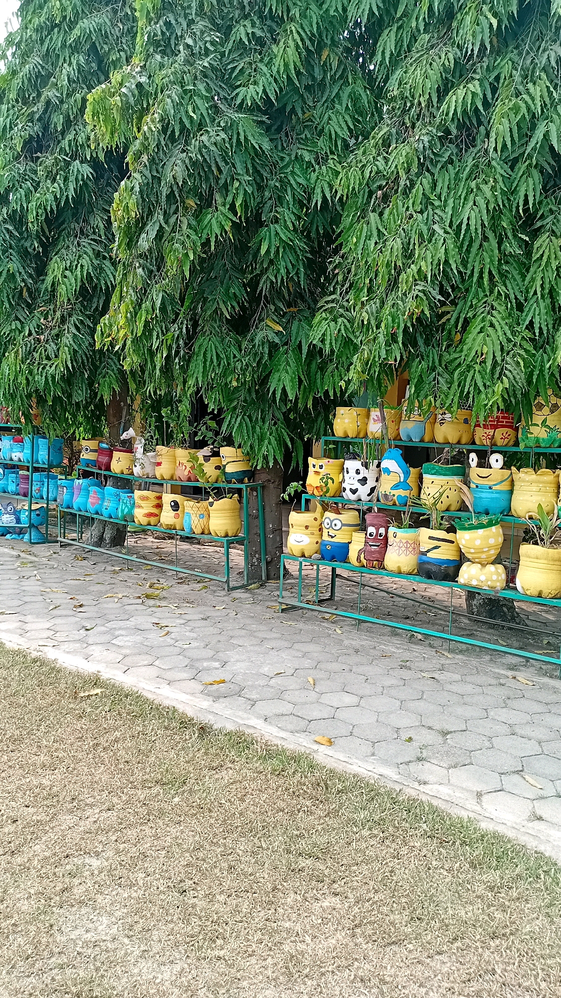 The image depicts a row of colorful, repurposed plastic bottles painted to resemble cartoon characters and animals, displayed on green metal shelves. These planters are arranged in an outdoor setting, with lush green trees providing a canopy overhead. The ground in front of the shelves is a paved walkway made of hexagonal stones, bordered by dry grass. The scene is likely located in Jambi, Indonesia, given the context. There are no people visible in the image. The planters are the central focus, showcasing creative reuse of materials and a playful aesthetic.