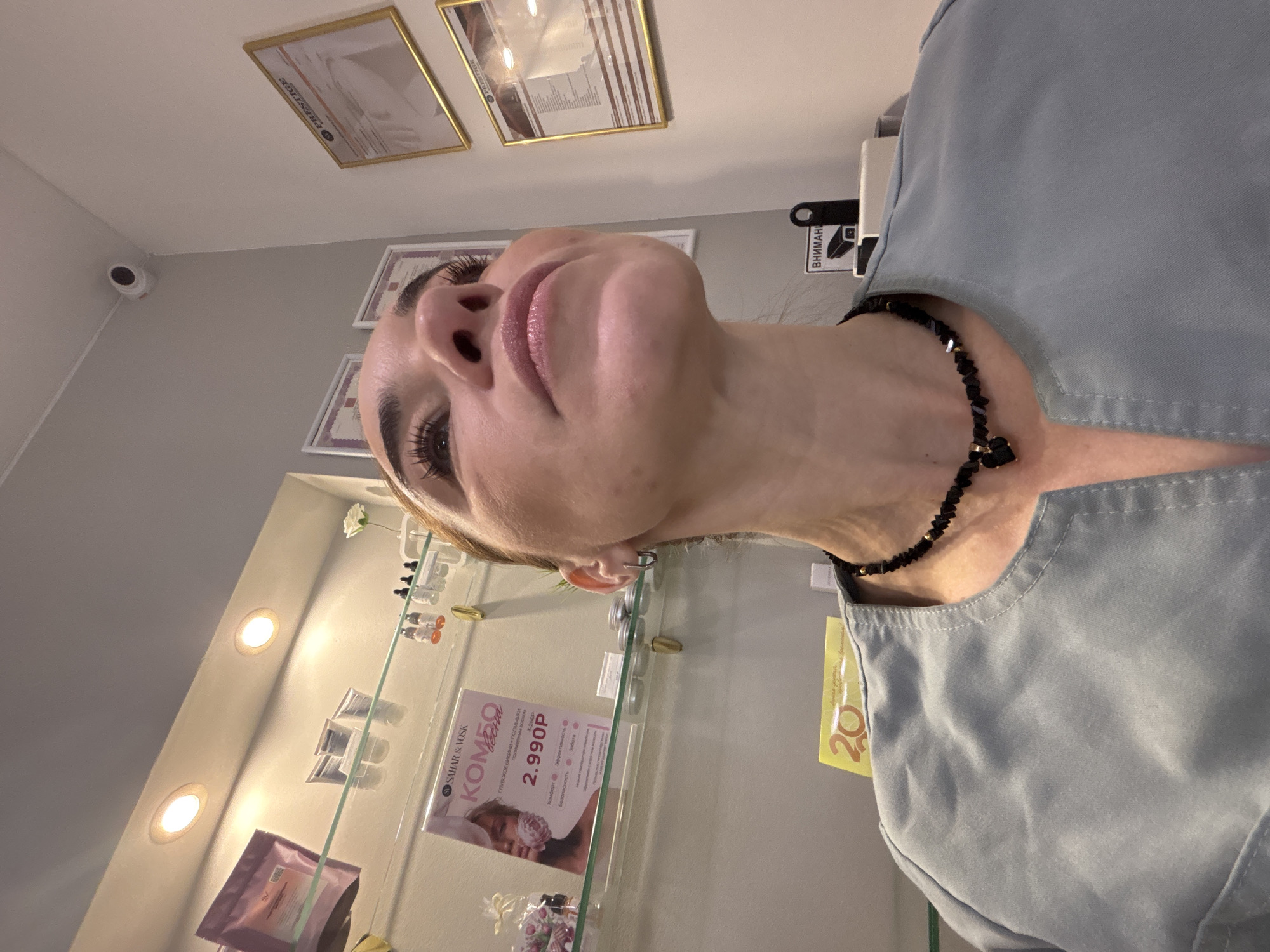 The image shows a close-up, low-angle view of a person's face and upper body. The person has fair skin, defined eyebrows, and long eyelashes. Their lips are pink and appear to have some gloss. They are wearing a black beaded necklace. The person appears to be in a beauty salon or similar establishment. In the background, there's a display case lit with small lights, showcasing various beauty products. One of the products is marked with "SAHAR & VOSK" and "KOMEO" and the price "2.990P." There are framed certificates on the wall in the background. A security camera is installed near the ceiling. A price tag with "20" printed on it is visible. Based on the product description in Russian, the establishment is possibly located in Vsevolozhsk, Russia.
