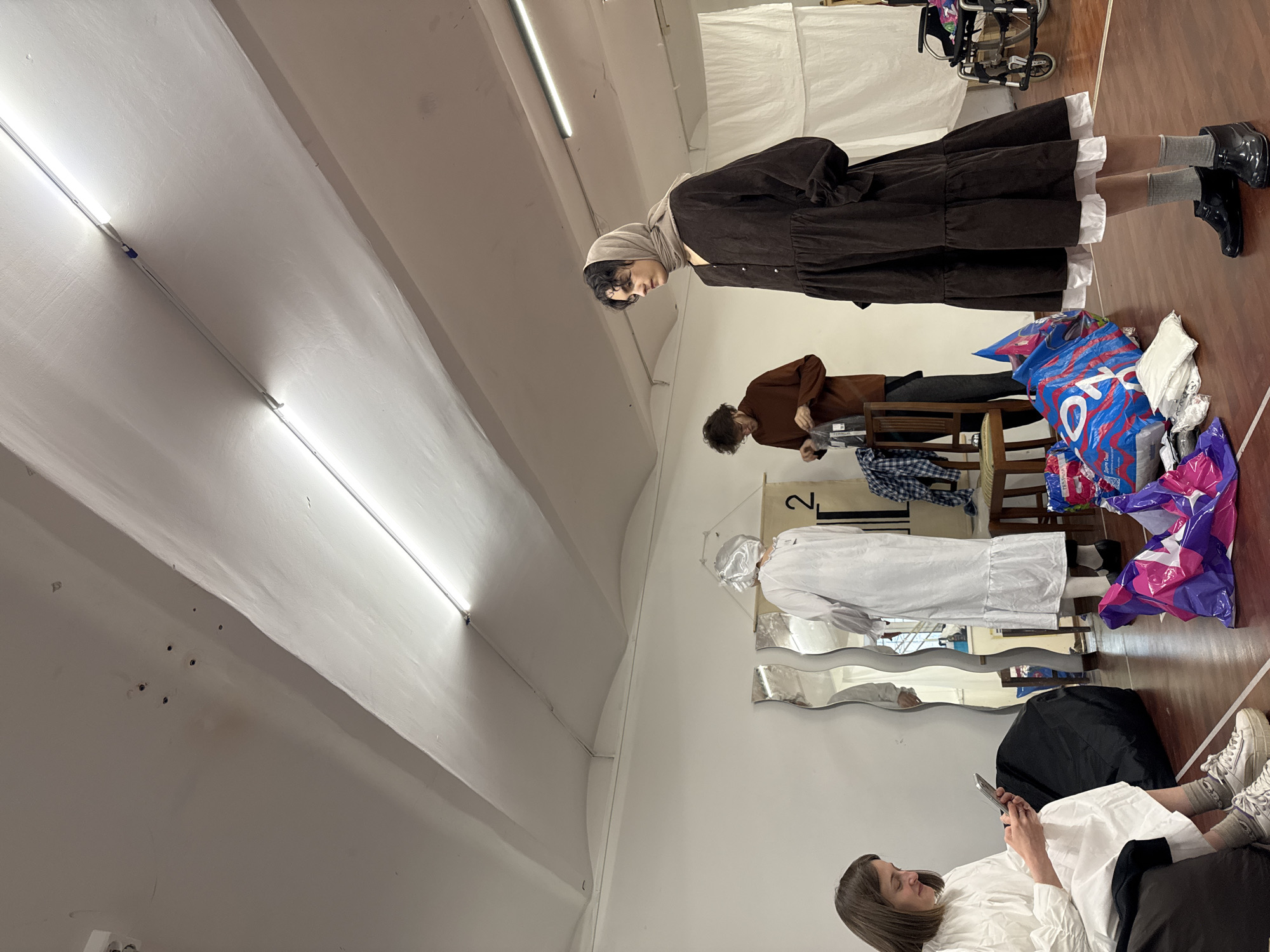 The image shows an interior space, likely a room used for fashion-related activities. Three people are visible. A person wearing a brown dress with a hijab stands near a wheelchair and shopping bags. Another person, in a brown shirt, stands near a chair. A third person, in a white dress, sits on a chair and looks at a mobile phone. A mirror on the wall is next to the sitting person. Fluorescent lights illuminate the room. The location, Moscow, Russia, is not evident from the image.