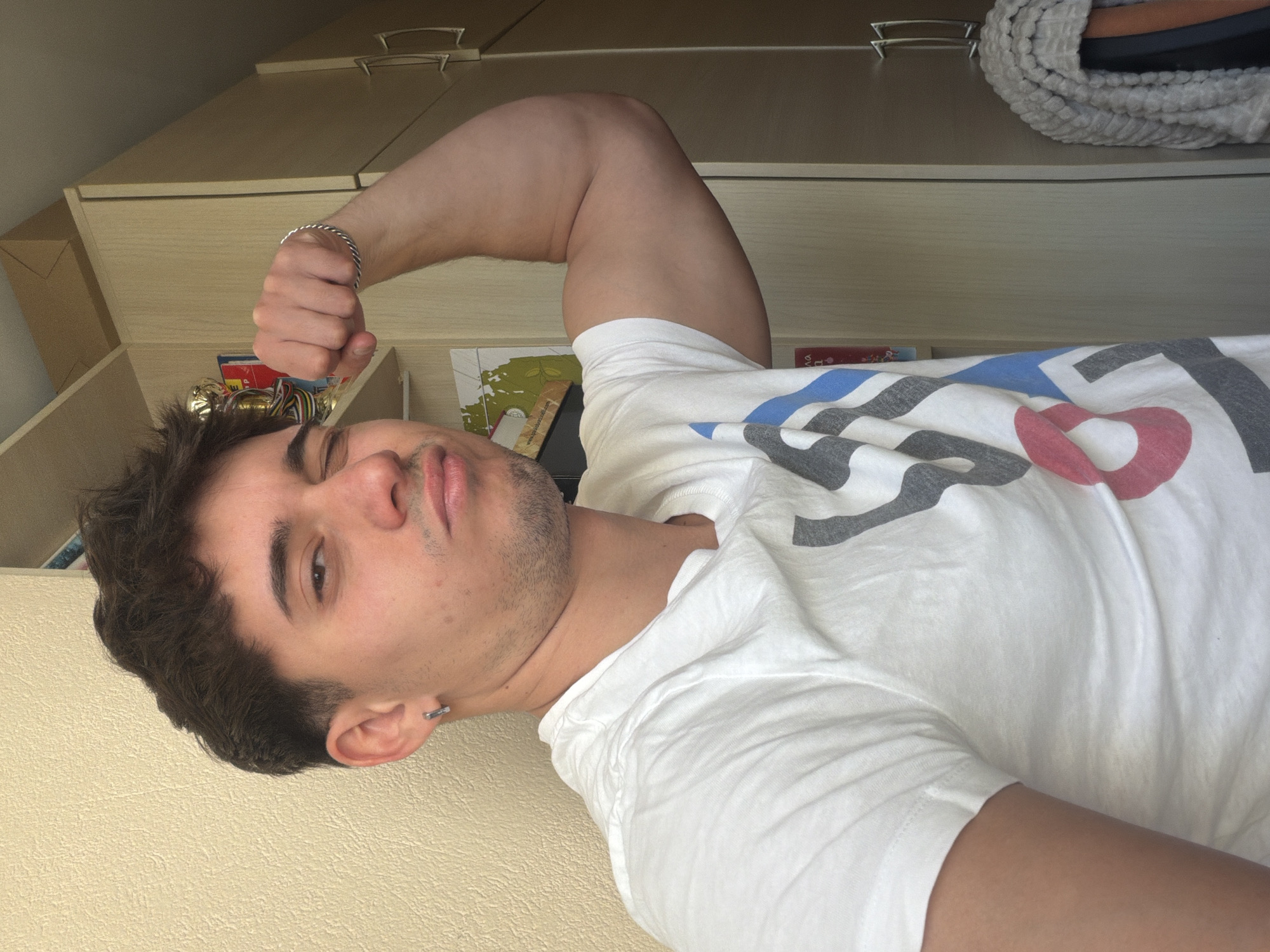 A light-skinned male with short, dark brown hair is pictured flexing his bicep. He is wearing a white t-shirt with a red, grey, and blue graphic design. He is also wearing a silver bracelet on his left wrist and a silver earring in his left ear. The background includes a light-colored wall, and a light brown wooden cabinet. Various small objects are visible on a shelf beneath the cabinet. The picture was most likely taken indoors. There is no clear evidence to determine if the image was taken in Blagoevgrad, Bulgaria.