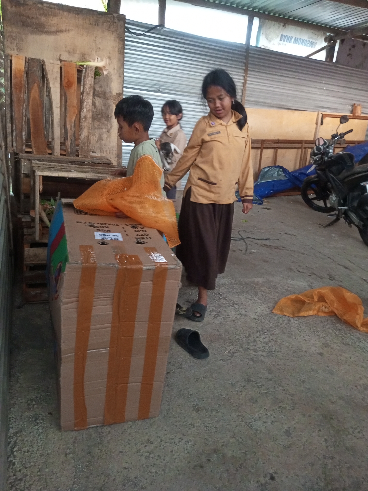 In Andongsili, Indonesia, three children are near a large cardboard box. A young girl in a tan uniform with a dark skirt stands over the box, looking down. To her left, a younger boy in a light green shirt watches something to his right. Behind them, another young girl, partially obscured, appears to be holding a small object. The box is taped with orange packing tape. An orange mesh bag rests on top of the box, and another similar bag lies on the ground nearby. A motorcycle is visible to the right, and the environment suggests an outdoor, possibly storage or work area, with corrugated metal roofing above.