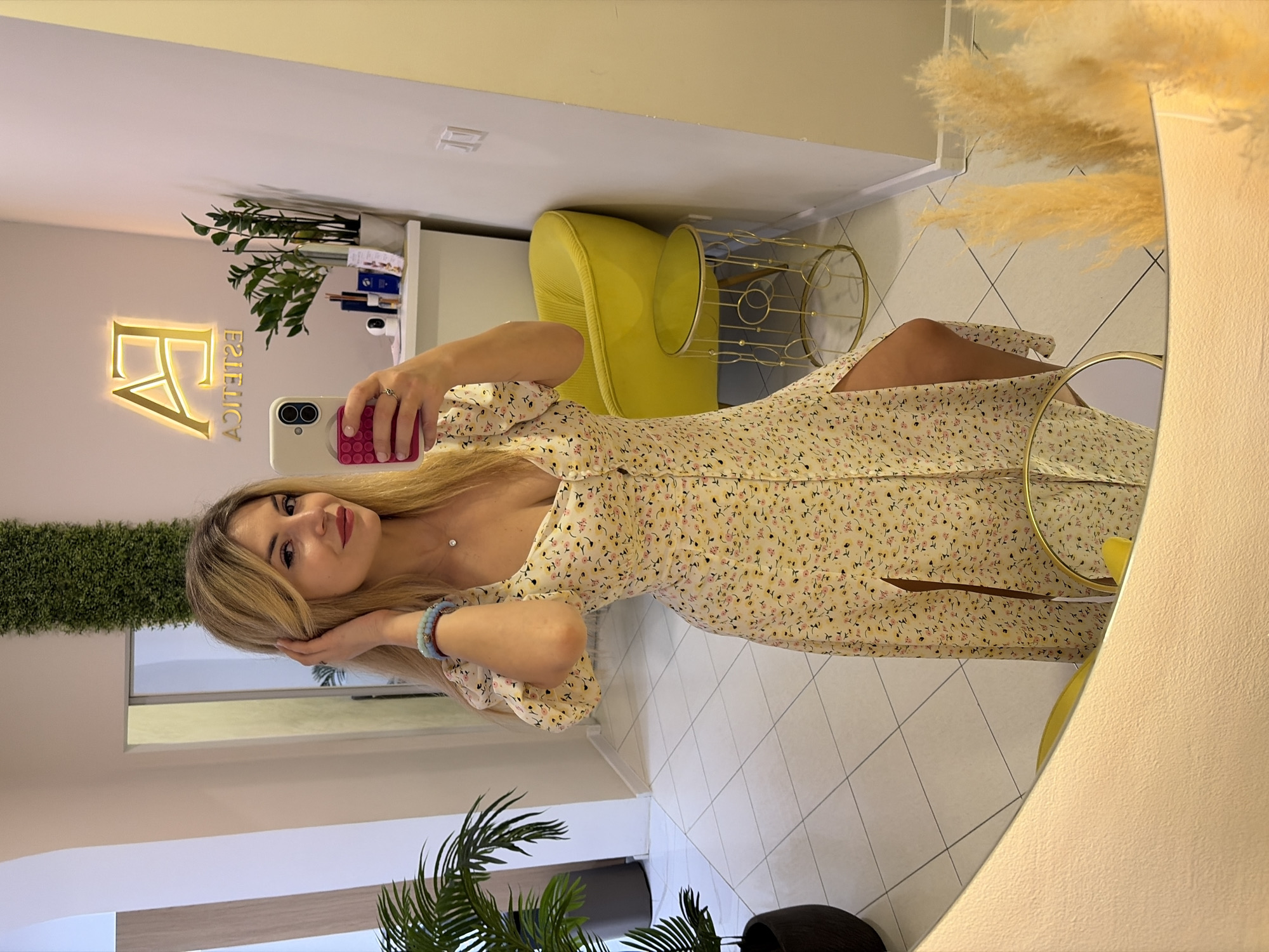 A young woman with long blonde hair, wearing a light-colored floral midi dress, takes a mirror selfie. She holds a white smartphone with a pink popsocket with her right hand, while her left hand gently touches her hair. She looks directly into the camera with a subtle smile. She wears a delicate necklace with a small pendant and light blue beaded bracelets on her left wrist.
The image is a reflection in a large, gold-framed mirror, revealing the stylish interior of what appears to be an aesthetic clinic or salon. On the wall behind her reflection, a glowing golden sign reads "FA ESTETICA" (mirrored). The reflected background also includes a bright yellow upholstered armchair, a golden wire-frame side table, a shelf with a potted plant, and decorative pampas grass. Directly visible parts of the room include a green, possibly artificial, plant panel on the left and large potted palm-like plants in the foreground corners. The floor is tiled with light-colored square tiles arranged in a diagonal pattern. The setting is indoors, well-lit, and presents a clean, modern aesthetic, consistent with a beauty establishment in Budva, Montenegro.