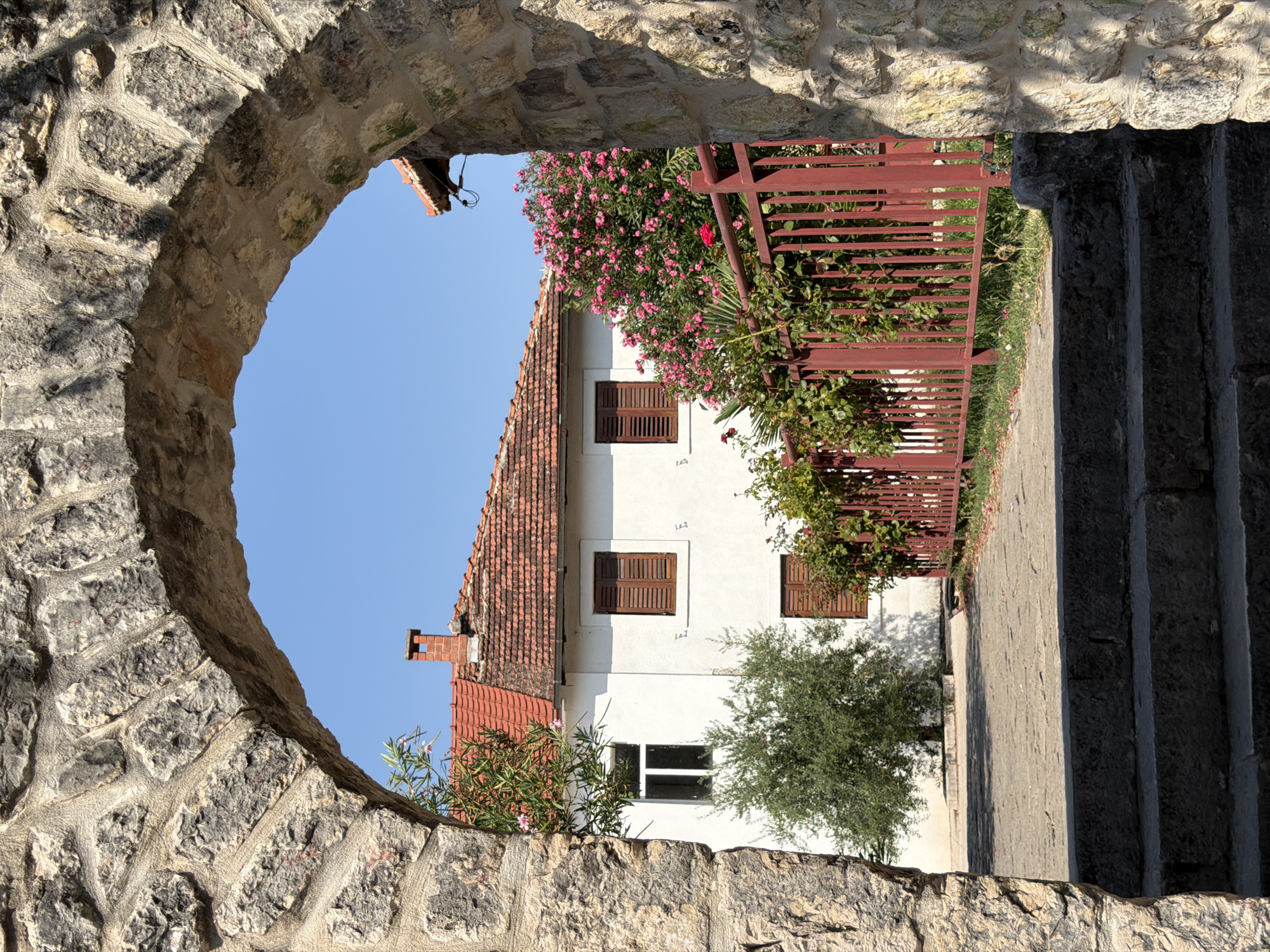 An outdoor, eye-level shot frames a white house with a red tile roof and brown shutters through a stone archway. The house has two visible windows on the front facade, one with white frames and the other with brown shutters. A small chimney with a red cap rises from the roof. To the right of the house, a dense bush with vibrant pink flowers stands behind a red metal fence. A small, green tree is visible to the left of the house. The ground in front of the house is paved with cobblestones. In the foreground, three rough-hewn stone steps lead up to the paved area. The scene is bathed in bright sunlight, casting shadows. The stone archway is made of large, irregular stones cemented together, with vegetation growing in some of the crevices. The sky is clear and blue. This appears to be a residential area in Trebinje, Bosnia and Herzegovina, possibly near the Old Town.