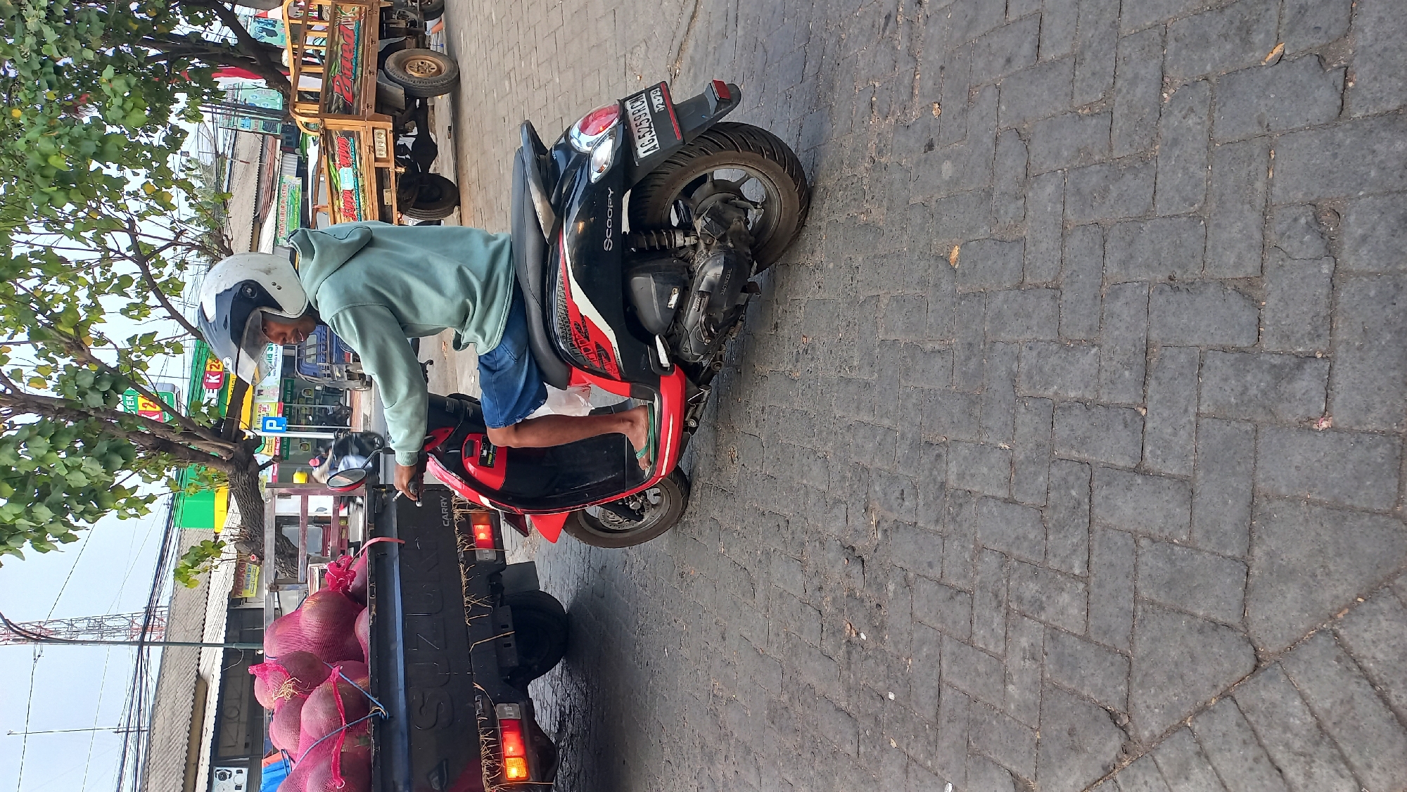 In Mangunsari, Indonesia, a person wearing a light blue hoodie and blue shorts is riding a red and black Honda Scoopy scooter. The rider has a white and black helmet on. The scooter is on a cobblestone street. To the left of the scooter, a black Suzuki Carry truck is visible, loaded with large, netted, red produce, possibly beets or yams. In the background, there are buildings, trees, and other vehicles, including a wooden trailer truck. The scene appears to be a busy street or market area.