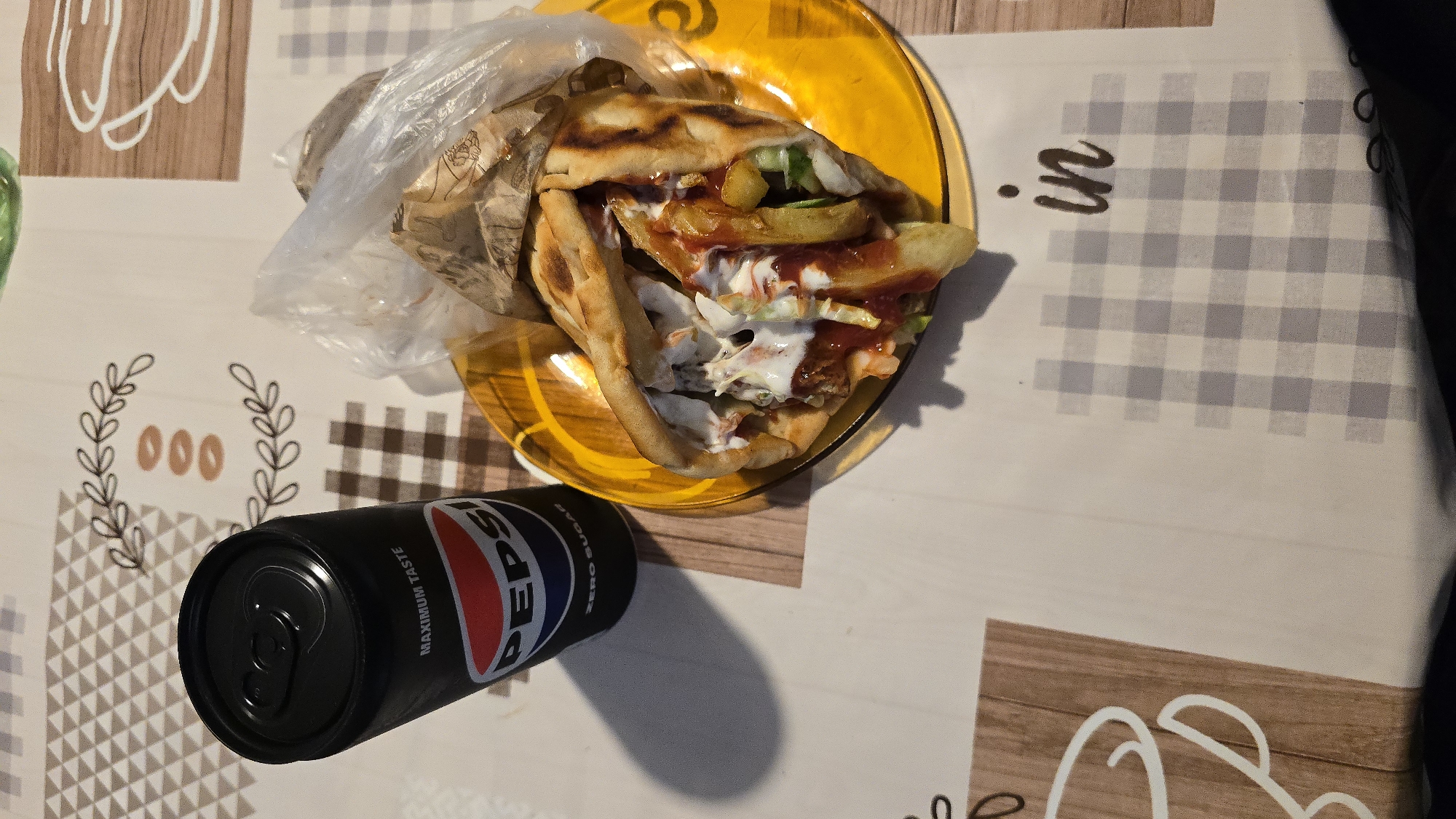 A black can of Pepsi Zero Sugar sits next to a gyro on a yellow plate, both resting on a checkered tablecloth. The gyro, a pita stuffed with meat, fries, lettuce, and sauce, is partially wrapped in paper and plastic. The tablecloth is white with brown and beige checkered patterns and decorative sprigs of leaves. The image is taken from a top-down perspective.