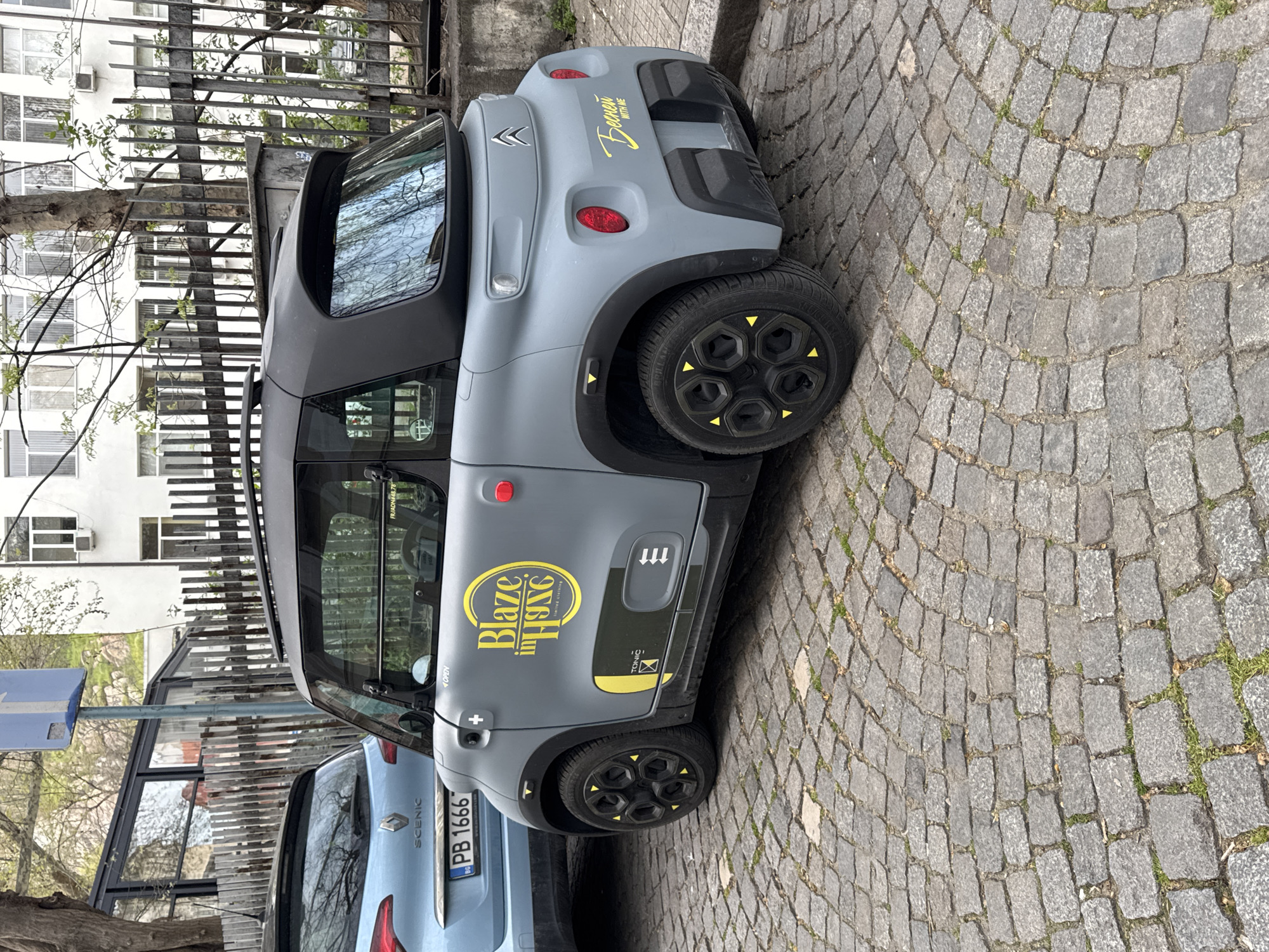 The image shows a gray Citroen Ami parked on a cobblestone street in Plovdiv, Bulgaria. The car has distinctive yellow accents on its wheels and side graphics that read "Blaze in HING" and "TONIC," along with a license plate that reads "PB 1666" and a "бесней WITH ME" text on the rear. The environment includes a metal fence, a building in the background, a Renault Scenic parked beside the Citroen Ami, and various signs. The sky is not visible.