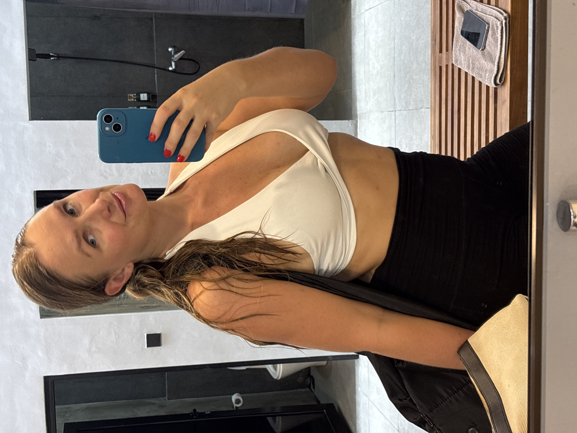 A woman is taking a selfie in a bathroom, likely in Kuta Selatan, Indonesia, based on the surroundings. She is wearing a white top and black bottoms. Her hair is wet. She is holding a blue iPhone with red nail polish on her fingers. A phone and a towel sit on a wooden shelf. The bathroom has gray and white tiles and dark fixtures. A black garment hangs partially visible over her shoulder.