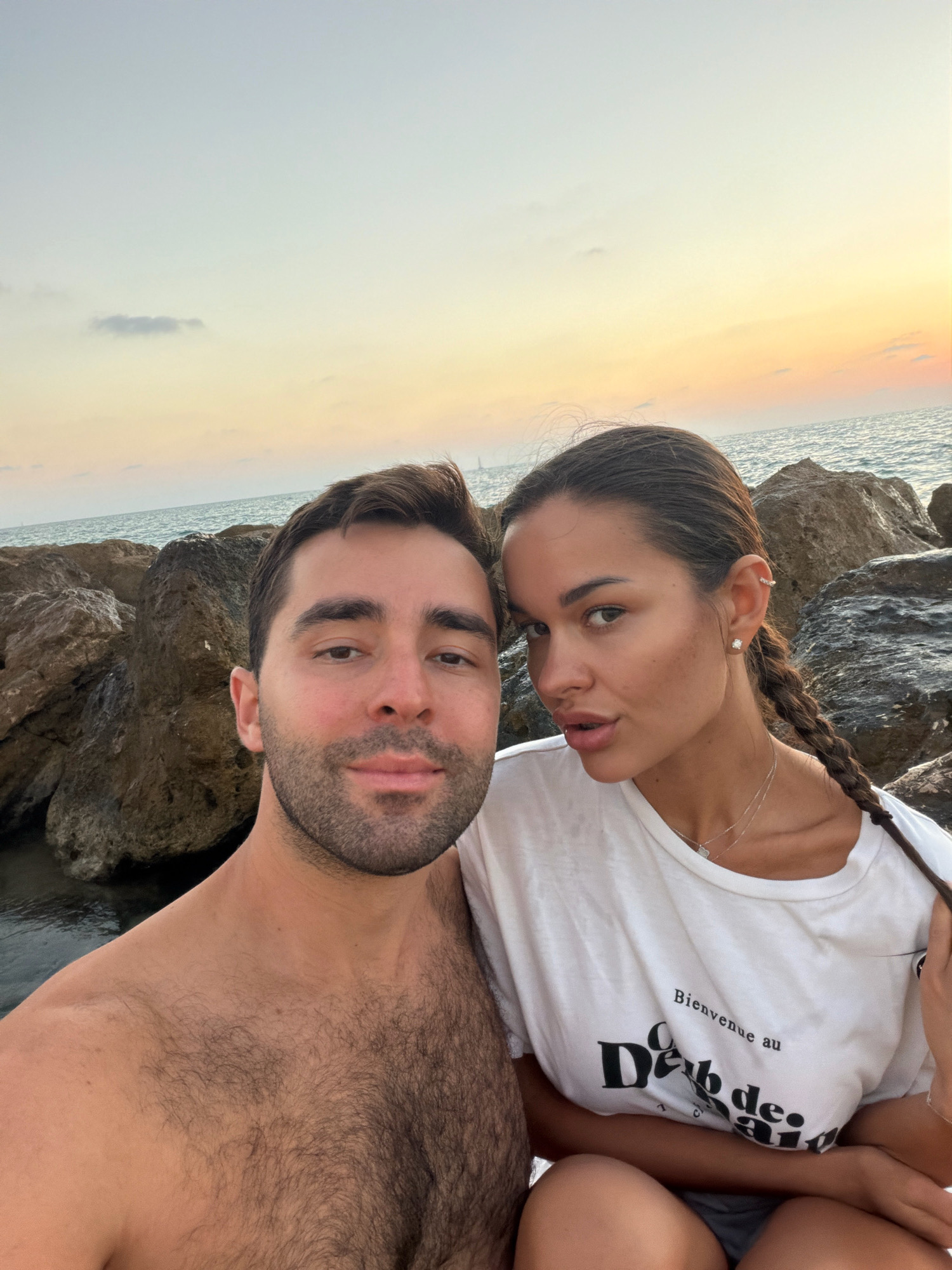 A selfie taken at dusk features a man and a woman posing in front of the sea. The man, shirtless and with a beard, is on the left, while the woman, wearing a white t-shirt with text, is on the right. They are seated on large rocks, likely on a beach. The ocean is visible in the background, with a soft, pastel-colored sky suggesting sunset or sunrise. The man's right shoulder and chest are prominently displayed, showing dark hair. The woman has a braid extending down her back and is wearing a necklace and earrings. The text on her shirt reads "Bienvenue au Deb de." The scene captures a romantic or casual moment by the sea. The location is likely Tel Aviv, Israel, given the context.