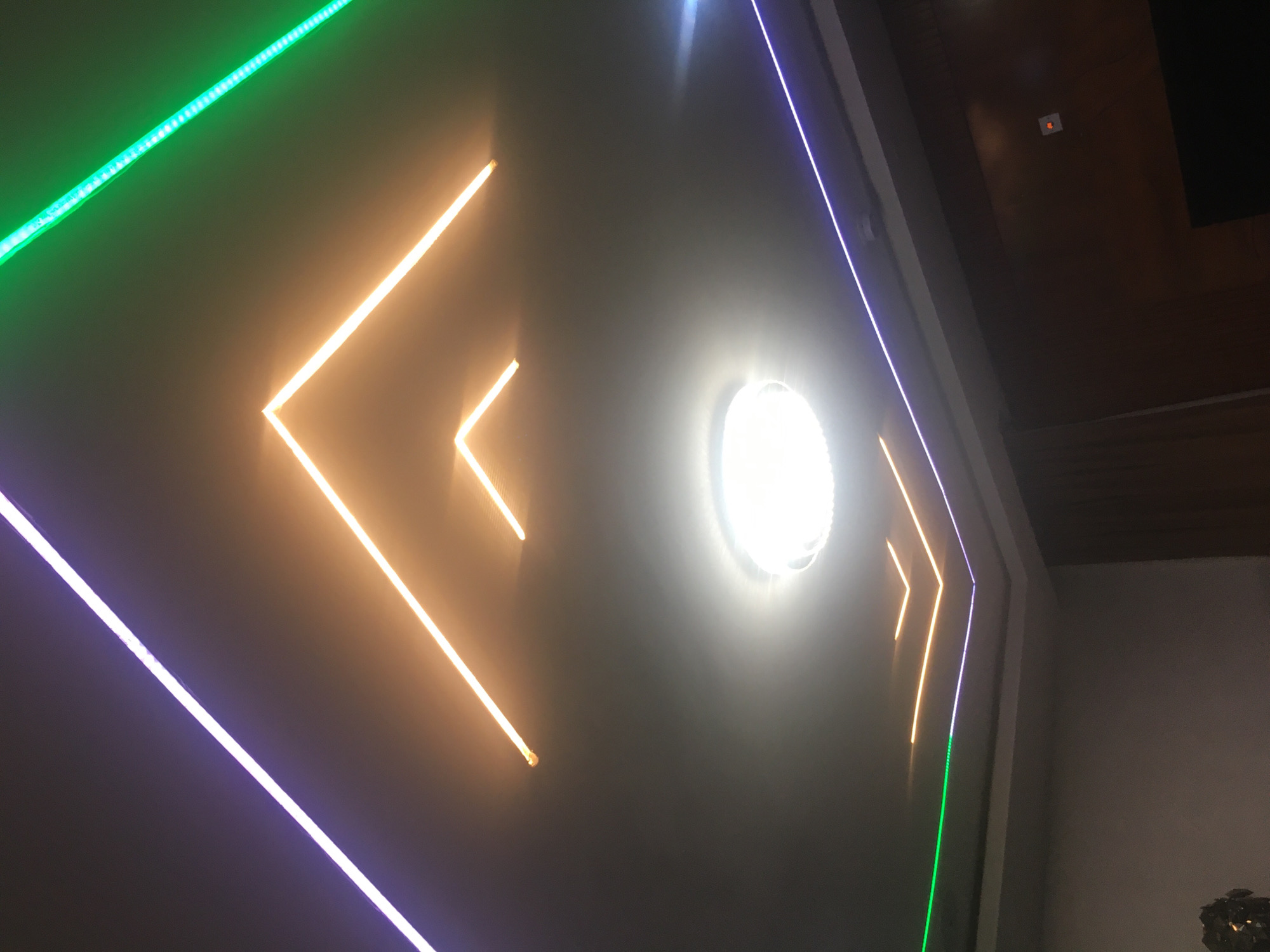 A low-angle shot depicts a gray wall illuminated by neon lights of green, orange, and purple. The lights form chevron-like designs on the wall. A bright, circular light fixture is also present. In the upper right, there is a glimpse of a dark brown ceiling, and a section of a lighter-colored wall is visible in the lower right. The location of the image is Jos, Nigeria, based on additional information not included in the image.