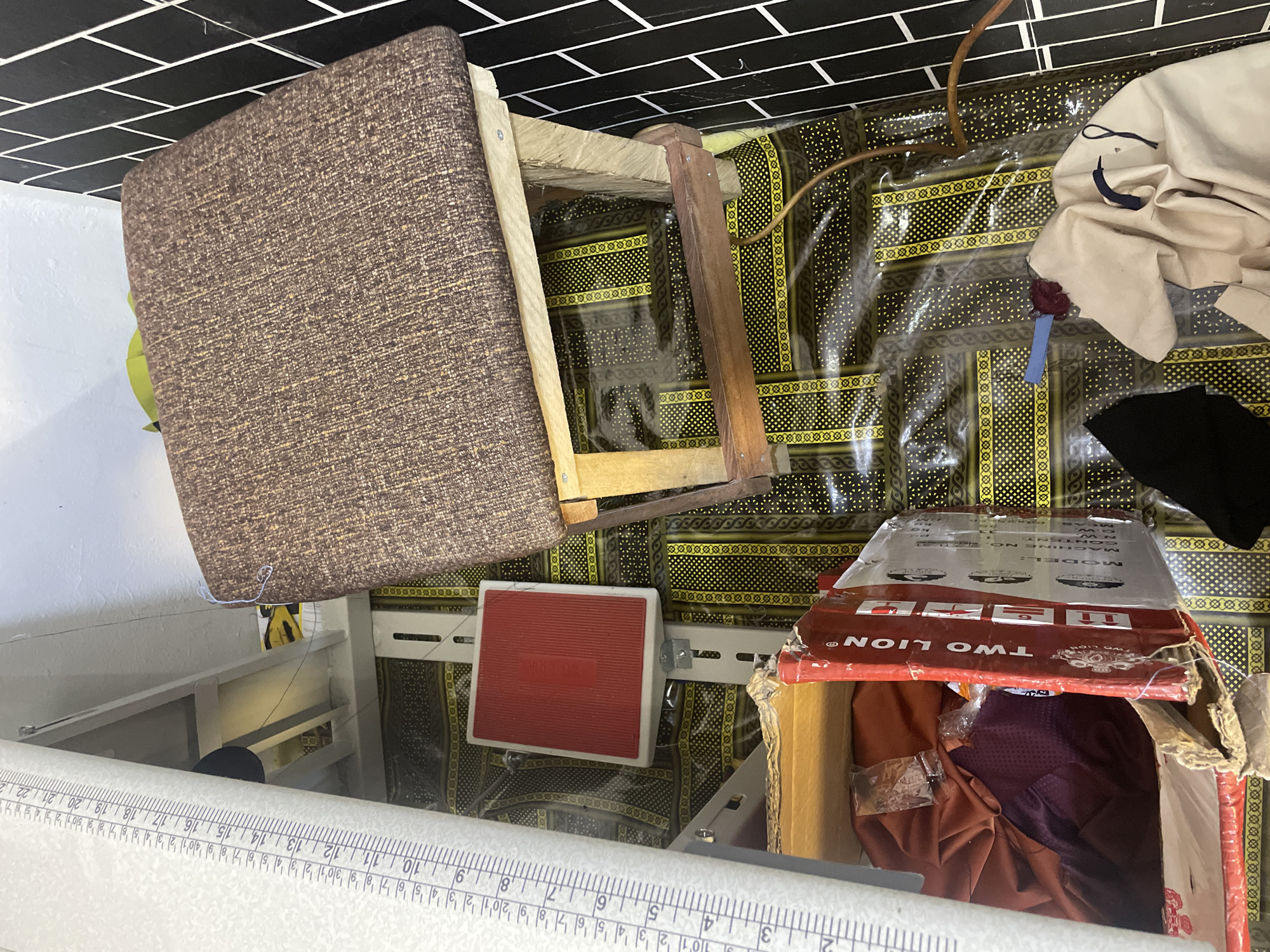 The image depicts an indoor scene, likely a workshop or storage area, with a focus on textile materials and sewing-related items. A large, white measuring tape with black markings is prominently displayed on the left side of the frame, indicating its use in measurements. In the center, a wooden stool with a brown, textured upholstered seat is positioned. Beneath the stool, a red and white object, possibly a foot pedal or a small platform, is attached to a grey metal rail. The floor is covered with a patterned material, featuring yellow and black geometric designs within clear plastic sheeting. To the right, a section of a black and white tiled wall is visible. In the foreground, a cardboard box labeled "TWO LION" is partially open, revealing folded fabric in shades of red and purple. Other fabric items and a dark piece of clothing are also present in the scene. The lighting suggests an indoor environment, possibly with artificial light. The specific location in Jalingo, Nigeria, is not determinable from the visual information provided.