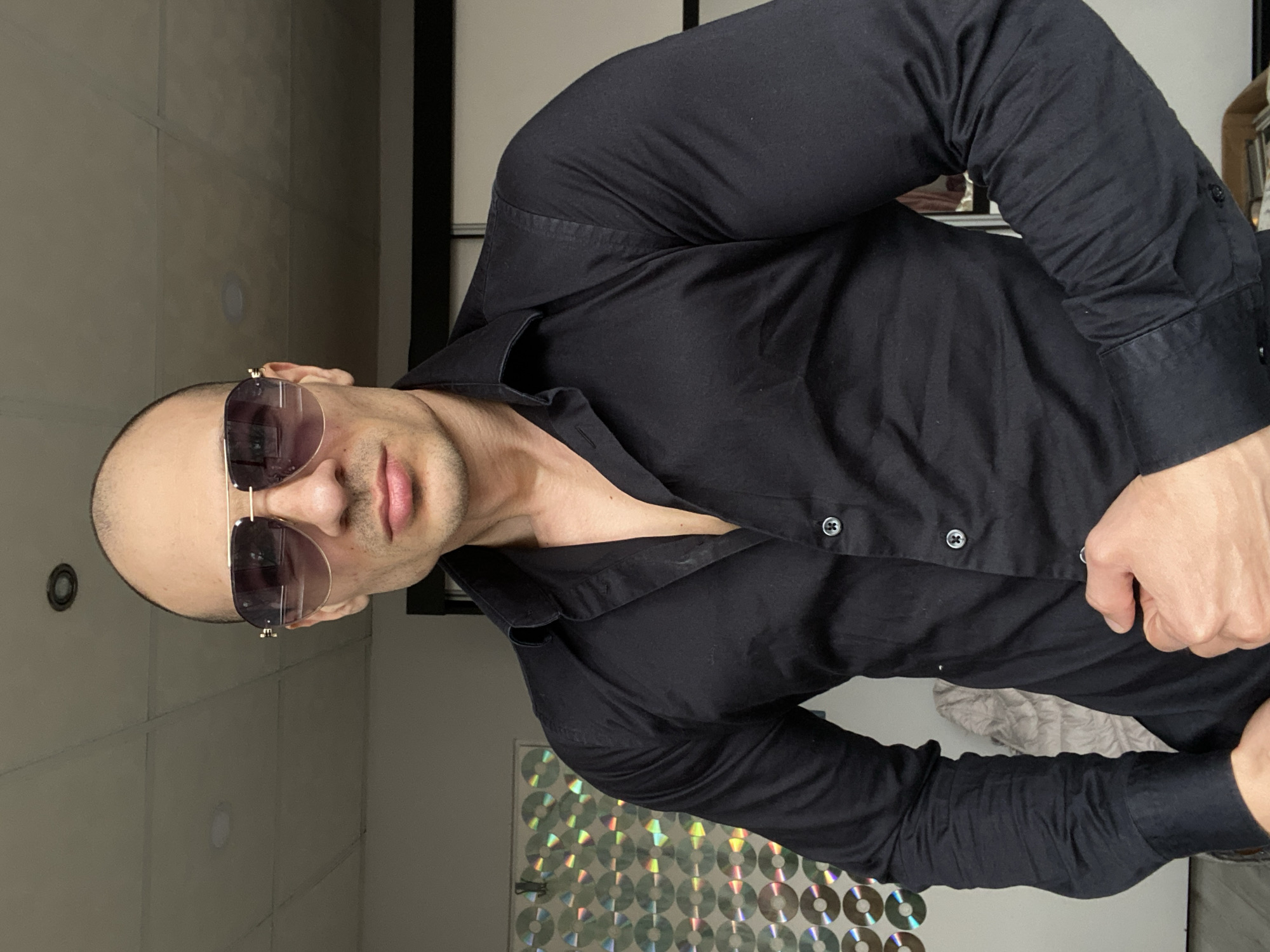 A man with a shaved head and sunglasses is posing indoors. He is wearing a black collared shirt and is standing near a wall covered in CDs. The location is not specified in the image and there is no indication it is in Pleven, Bulgaria.