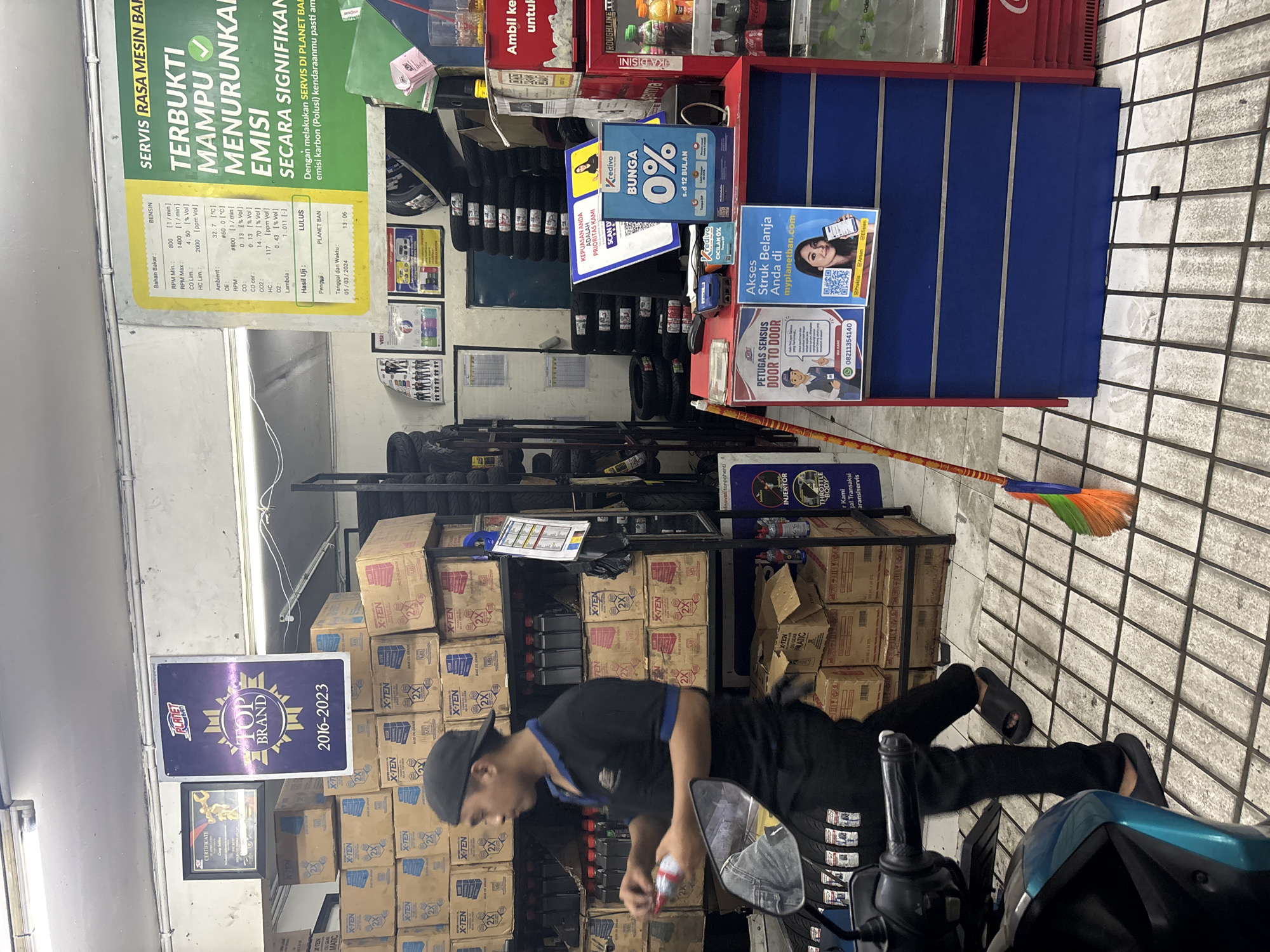 The image shows the interior of a motor vehicle service shop in North Jakarta, Indonesia. In the foreground, a man wearing a black t-shirt, black pants, and black slippers is holding a red and white bottle, appearing to be pouring its contents. He is standing near a motorcycle. Behind him, shelves are stacked with boxes labeled "X-TEN" and "MB," presumably containing engine oil or other automotive fluids. A broom with colorful bristles leans against a shelf.
In the background, a large green sign with white text reads "SERVIS RASA MESIN BARU TERBUKTI MAMPU MENURUNKAN EMISI SECARA SIGNIFIKAN". To the left of this sign is a white poster with a table of technical data related to engine performance, and above it, a framed "TOP BRAND 2016-2023" award.
Further to the right, a red counter displays various promotional materials. One prominent poster is blue and advertises "KREDIVO BUNGA 0% s.d 12 BULAN." Another poster shows a QR code and text related to "myplanetban.com". A small card reader is placed on the counter. A refrigerator unit with drinks is visible on the far right. The floor is tiled with a grid pattern. The overall environment suggests a busy service center.