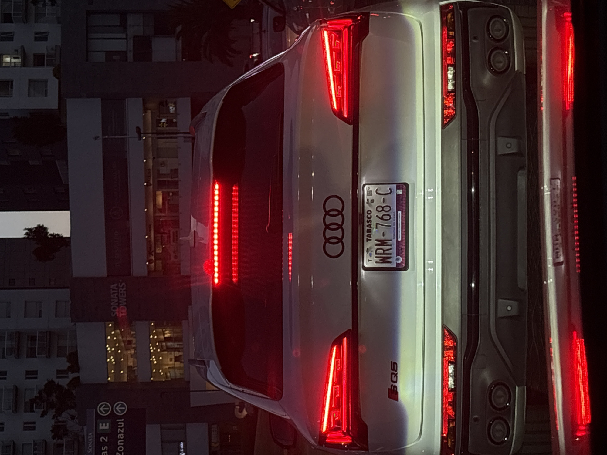 The image, rotated 90 degrees counter-clockwise, depicts the rear view of a light-colored (likely silver or white) Audi SQ5 at night, set in an urban environment, likely San Bernardino Tlaxcalancingo, Mexico. The car's prominent red LED taillights are illuminated, creating bright streaks of light. The Audi rings emblem is centrally placed on the tailgate, with the "SQ5" model designation visible on the lower right. A Mexican license plate from Tabasco state, bearing the characters "WRM-768-C", is clearly displayed. Dark-colored lower bumper elements, possibly housing exhaust outlets or a diffuser, are also visible.
In the dark background, several buildings are present, some with illuminated windows, indicating nighttime. Text visible on the buildings and signs includes "SONATA TOWERS", "SONATA", "Zonazul" with an upward arrow, and "RIVIERA". Another sign reads "as 2 E". Tree foliage is sparsely visible against the buildings. The scene suggests the vehicle is stationary, possibly in traffic or parked, within a developed city area. Another vehicle's red taillight is partially visible on the far right, suggesting other cars are nearby.