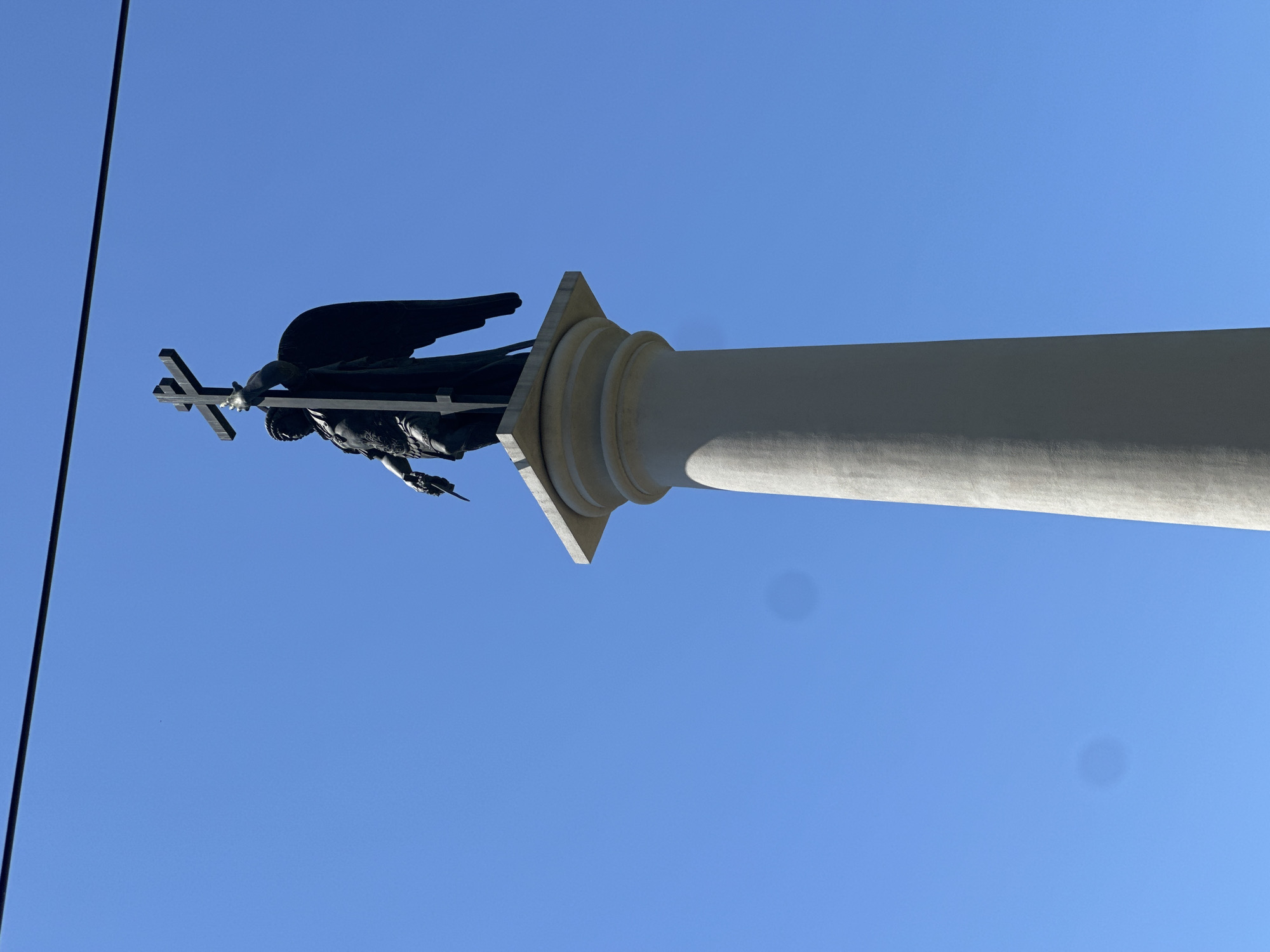 A dark statue of an angel holding a cross stands atop a tall, light-colored column against a clear blue sky. The statue is silhouetted and depicts the angel with wings, wearing armor and a laurel wreath. The column is cylindrical and tapers slightly towards the top, where it supports a square pedestal. A thin, dark wire or cable runs horizontally across the upper portion of the image, above the statue. The context provided indicates this is the monument to Archangel Michael in Sochi, Russia, symbolizing protection over the city.