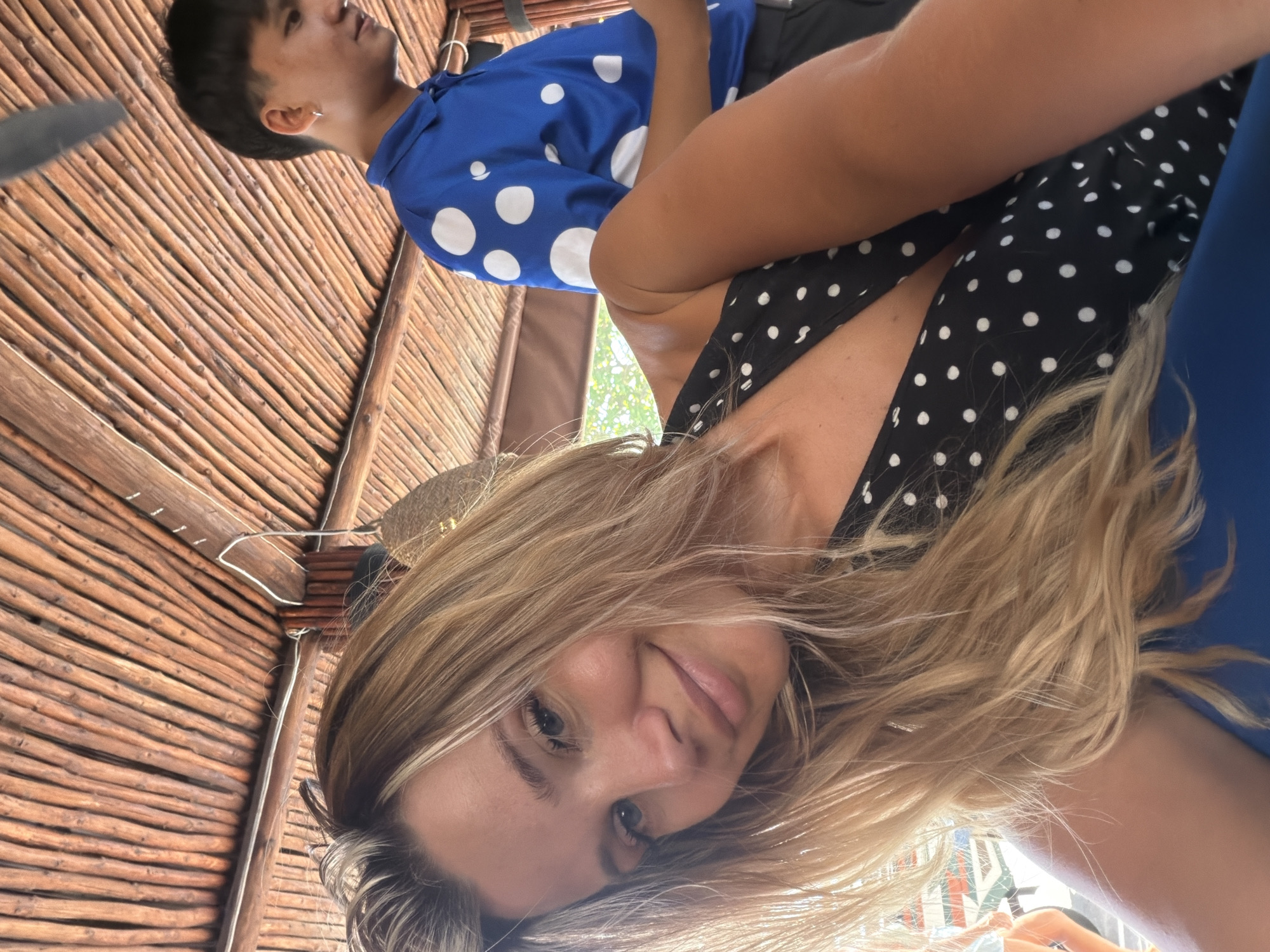 A woman with blonde hair smiles at the camera in a close-up selfie. She is wearing a black dress with white polka dots. Behind her, a young man in a blue polka-dotted shirt is visible. The background suggests an outdoor setting with a thatched roof and bamboo structures, indicative of a restaurant or bar. The lighting is bright, suggesting it is daytime. The scene appears to be in Karon, Thailand, given the style of architecture and the tropical vegetation visible in the background.