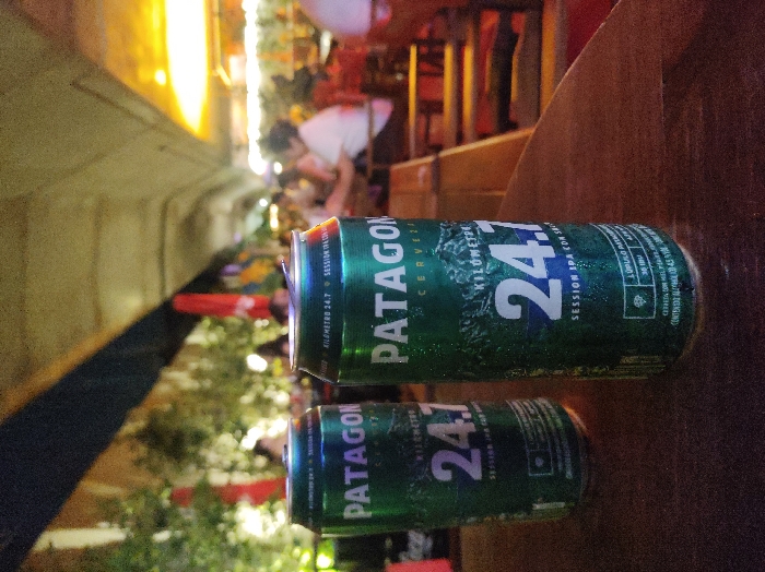 Two green Patagonian beer cans sit on a wooden surface. "PATAGONIA KILÓMETRO 24.7" is prominently displayed on each can. The upper can is larger and appears to be in focus. The background features an outdoor setting with blurred figures, trees, and colorful lights. The composition is shot at an angle, with the cans dominating the foreground.