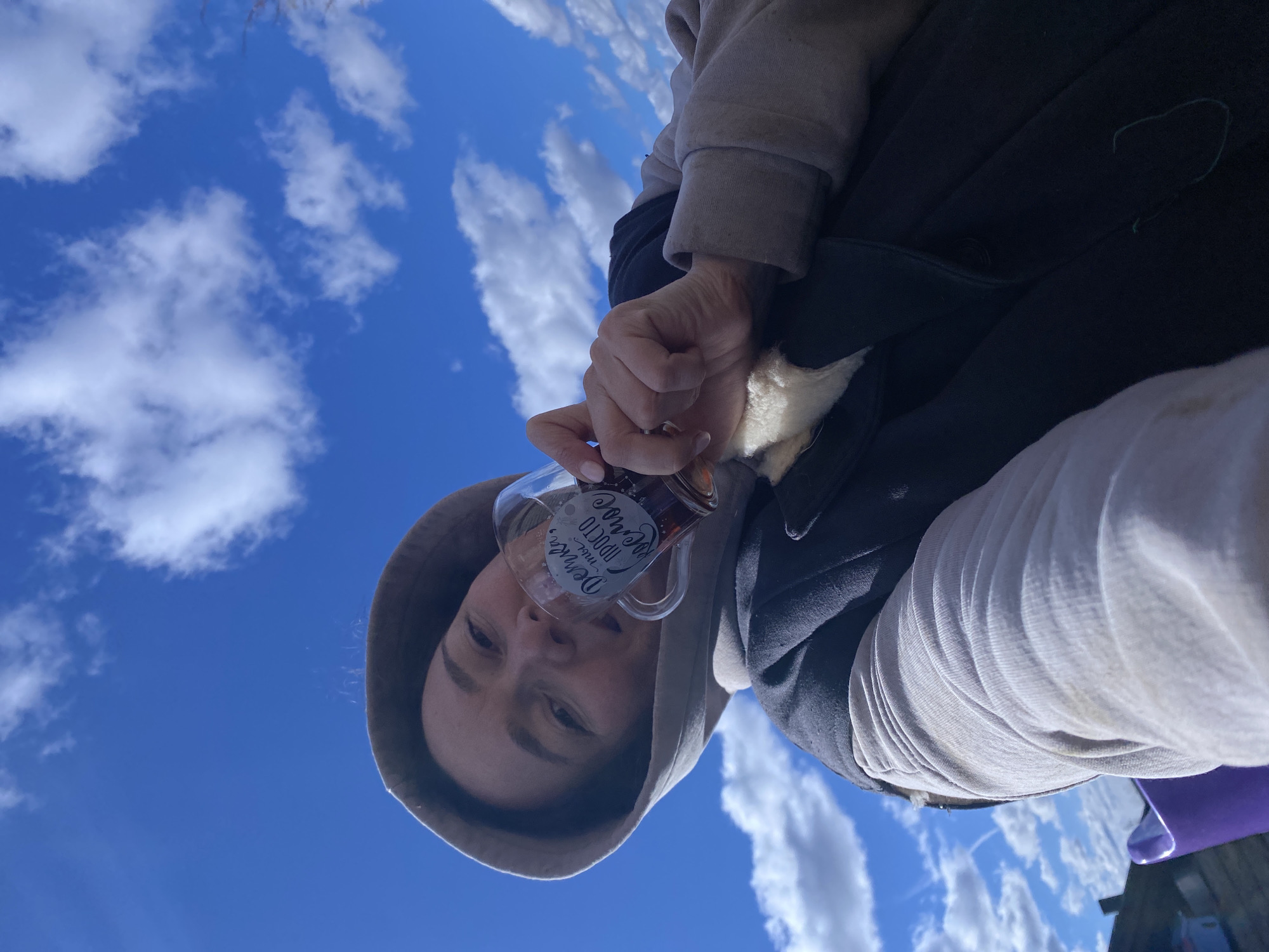 A low-angle shot shows a person wearing a hooded sweatshirt, holding a mug with lettering on it against a blue sky with scattered clouds. The person's face is visible inside the hood. They are wearing a jacket with a fur-like lining. The mug contains a dark liquid. The location is in the city of Ольховка, Russia.