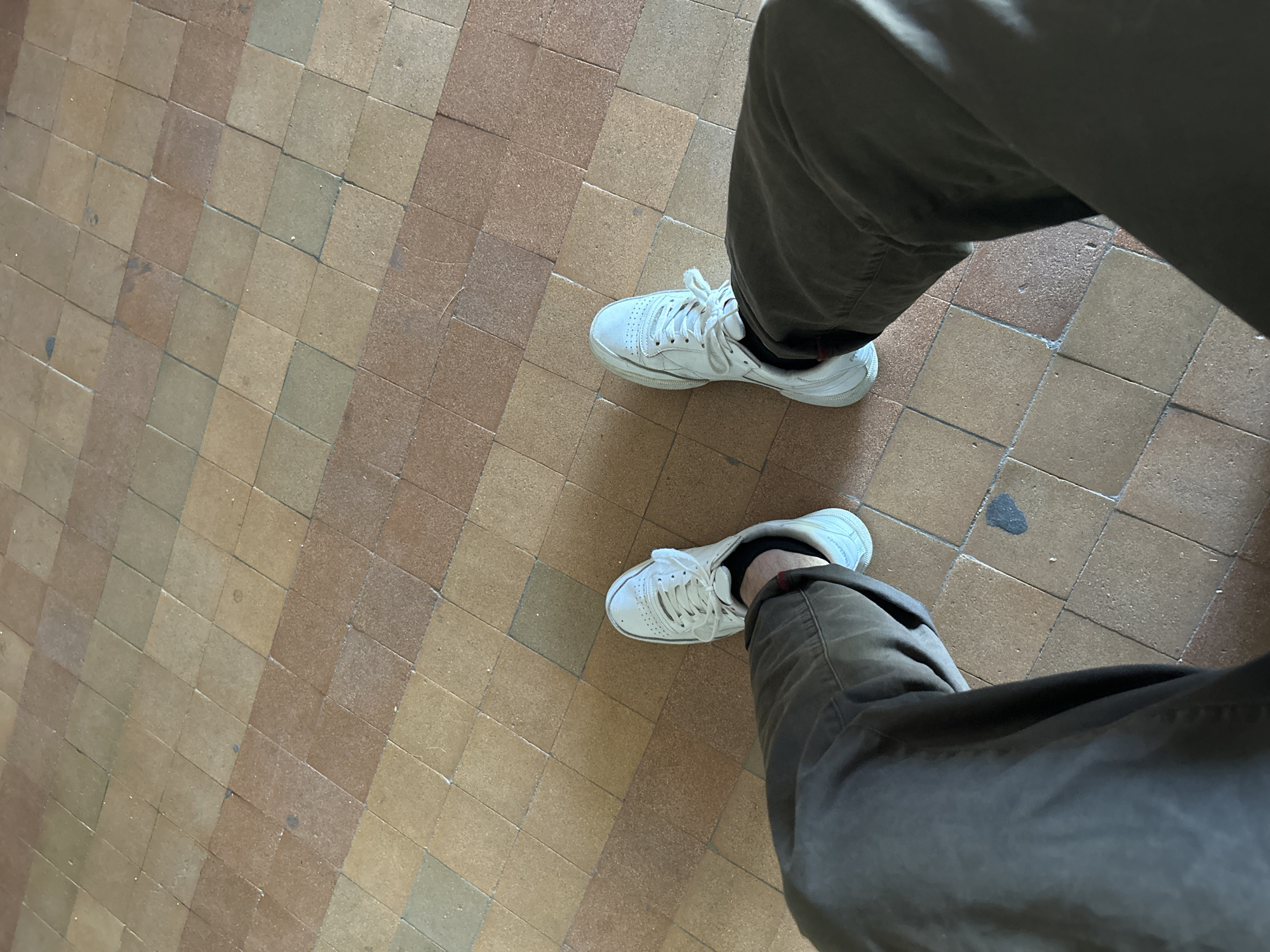The image shows a first-person perspective of a person's legs and feet standing on a tiled floor. The person is wearing dark-colored pants and white sneakers. The floor is composed of square tiles in varying shades of brown and tan, arranged in a checkerboard pattern. The scene is lit by overhead lighting, casting shadows on the floor. The image does not contain any identifiable landmarks or features that would indicate a specific location within the city of Slobozia, Moldova.
