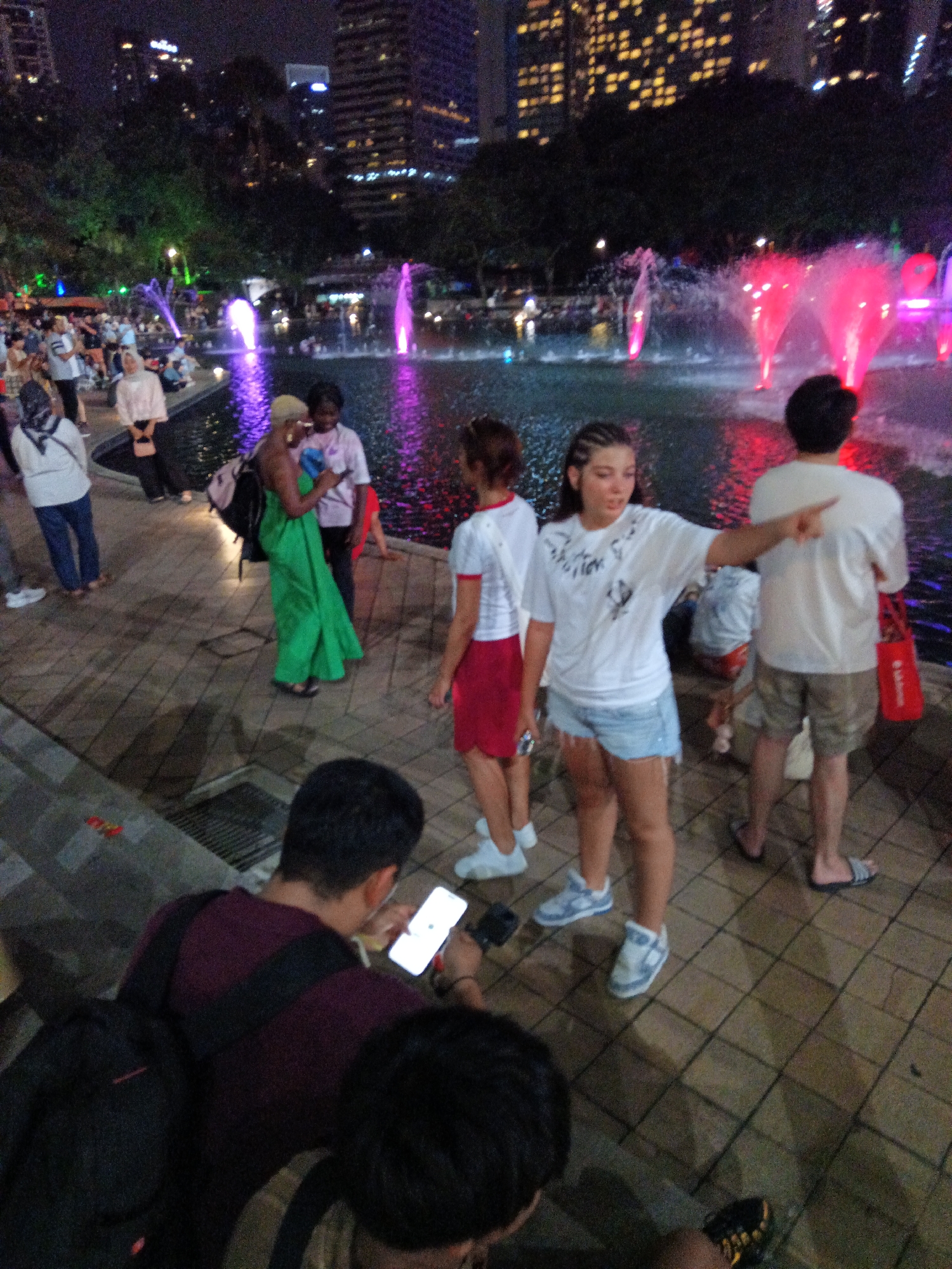 This image depicts a nighttime scene at the Kuala Lumpur City Centre Park, with the Petronas Twin Towers visible in the background. A group of people are gathered near a fountain illuminated with pink and purple lights. In the foreground, a person wearing a burgundy shirt and a backpack is looking at a smartphone. To their right, a young woman with braided hair is wearing a white t-shirt and denim shorts and is pointing to the right. Another woman in a green dress stands nearby, looking at her phone. Other individuals are seen in the mid-ground, some sitting and others standing, observing the fountains and the cityscape. The park features a paved walkway, trees, and a body of water.