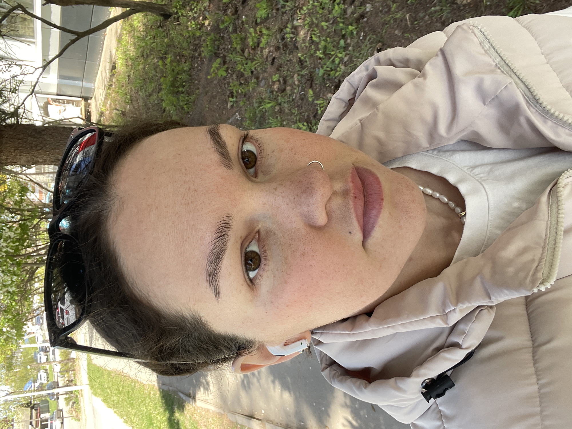 A close-up, slightly angled, daytime shot shows a woman in what appears to be an outdoor setting. She is wearing a light-colored, possibly beige, puffy jacket over a white shirt with a pearl necklace. The woman has fair skin, brown eyes, and dark hair, with sunglasses perched on her head. A small gold ring is visible in her nostril, and she is wearing an in-ear headphone.
The background includes greenery, suggesting a park or other outdoor space. It's difficult to determine the precise location in Izhevsk, Russia, without more context. The environment looks like a typical urban park or sidewalk area.