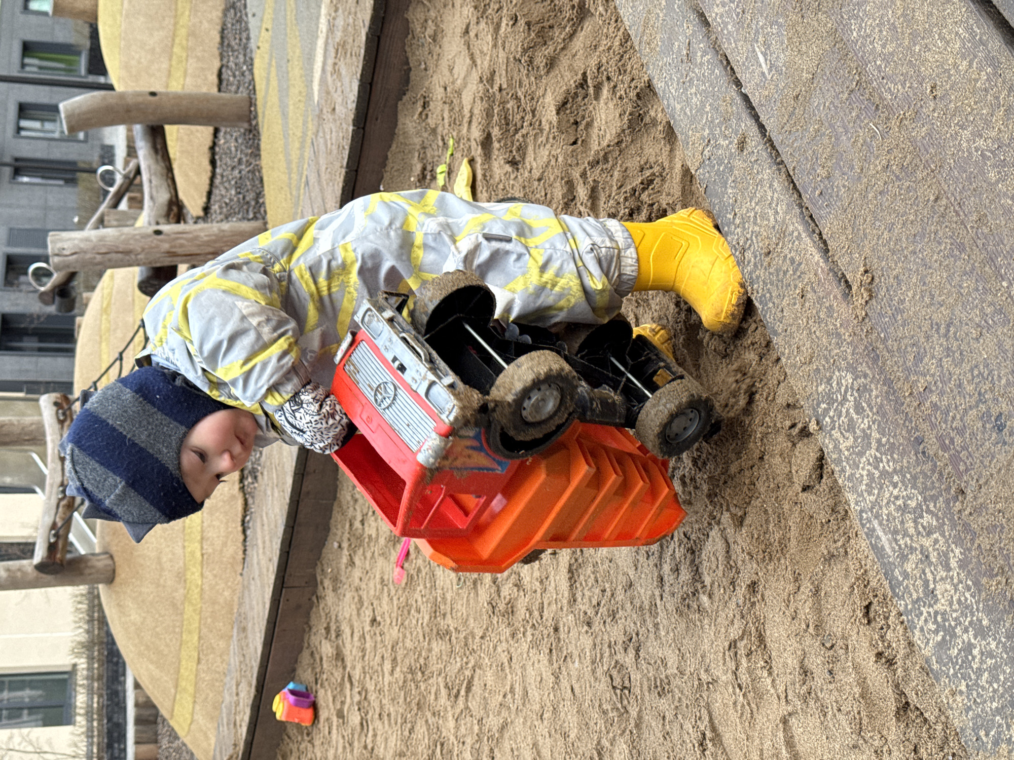 A young child, likely a toddler, is actively engaged in playing in a sandpit, presumably at a playground in Izhevsk, Russia. The child is focused on a large, red and orange toy dump truck, which is partially filled with sand, indicating active play.
The child is dressed in warm outdoor attire, consisting of a grey jacket and pants set adorned with a yellow abstract pattern, bright yellow rubber rain boots, and a striped blue and grey knit hat with small ear-like protrusions. Their face is partially visible, showing concentration on the toy.
The immediate environment is a sand-filled area bordered by dark wooden planks on one side. Various wooden playground structures, including posts and beams, are visible in the background, further confirming the playground setting. Beyond these structures, modern multi-story buildings with windows are visible, suggesting an urban or suburban location. A small, colorful toy is also present in the sand near the child's head.
The scene is well-lit by natural light, indicating it is daytime, possibly under clear skies. There is no visible text in the image. The overall impression is of a child enjoying a playful and engrossing outdoor activity.