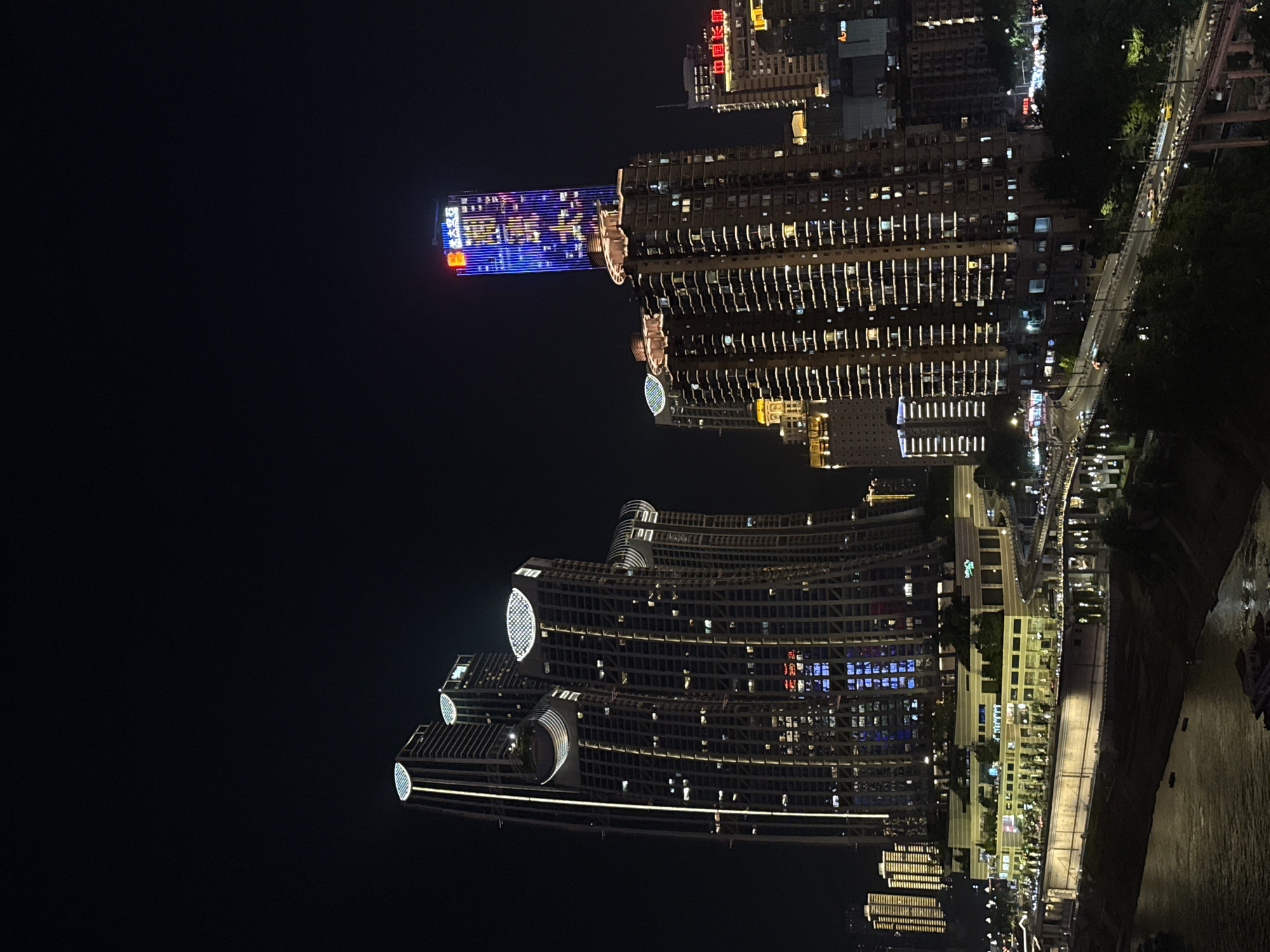 This image captures a nighttime view of the Yuzhong District in Chongqing, China, showcasing a dense urban landscape illuminated by artificial lights. Dominating the skyline are several modern skyscrapers. On the left, a cluster of tall buildings features curved facades and distinctive circular elements near their tops, illuminated by yellow and white lights. Some of these buildings have vertical lighting strips running along their edges.
To the right of this cluster, a prominent, slender skyscraper stands tall, adorned with a vibrant blue light display that includes some text and patterns. This skyscraper is surrounded by other buildings of varying heights, also lit up. One of these buildings displays red and yellow signage.
Below the main skyscrapers, a multi-tiered structure, possibly a shopping mall or commercial complex, is visible. It is illuminated with yellow and white lights, and trees and greenery are present on its terraces. A river flows in the foreground, reflecting the city lights. Elevated roadways and bridges, also lit, traverse the scene, with vehicles visible on them. The overall impression is one of a bustling, brightly lit metropolis at night, with a mix of architectural styles and a significant riverfront presence.