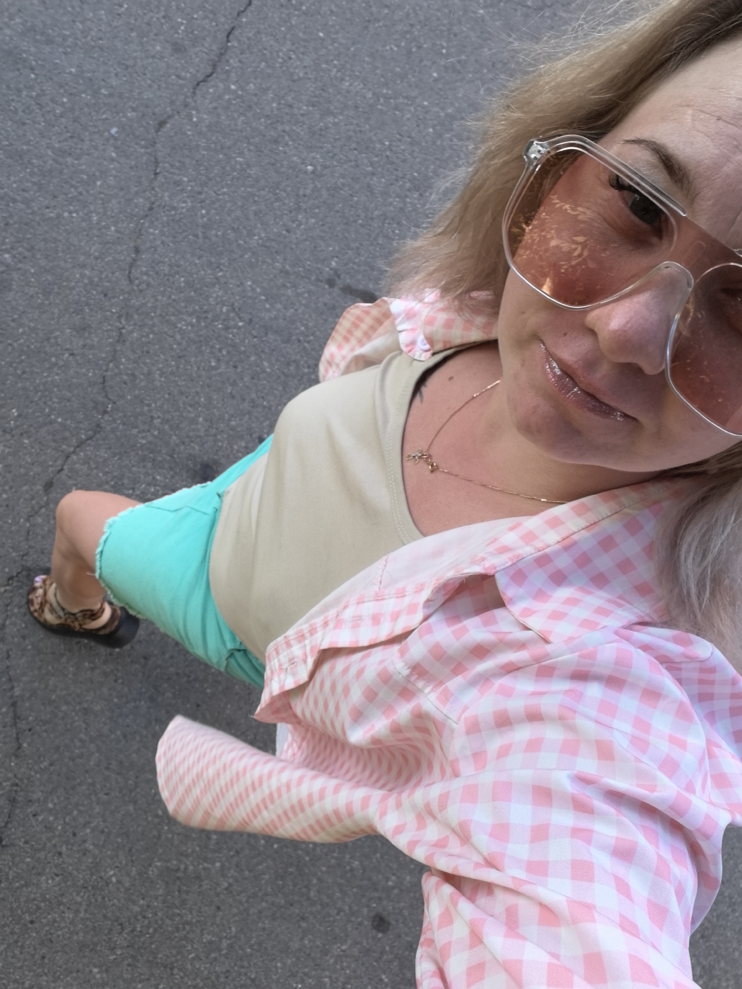 A woman is pictured from a high angle, standing on a cracked asphalt surface. She has fair skin and blonde hair. She is wearing large, rose-tinted, clear-rimmed sunglasses. Her lips are glossed and appear slightly parted. Around her neck, she wears a thin gold chain with a small pendant. She is dressed in a light tan t-shirt, turquoise shorts, and a pink and white gingham shirt, open at the front. She is also wearing brown and black patterned sandals. The scene appears to be outdoors in an urban environment, possibly Vityazevo, Russia, given the context of the request. The lighting suggests it is daytime.