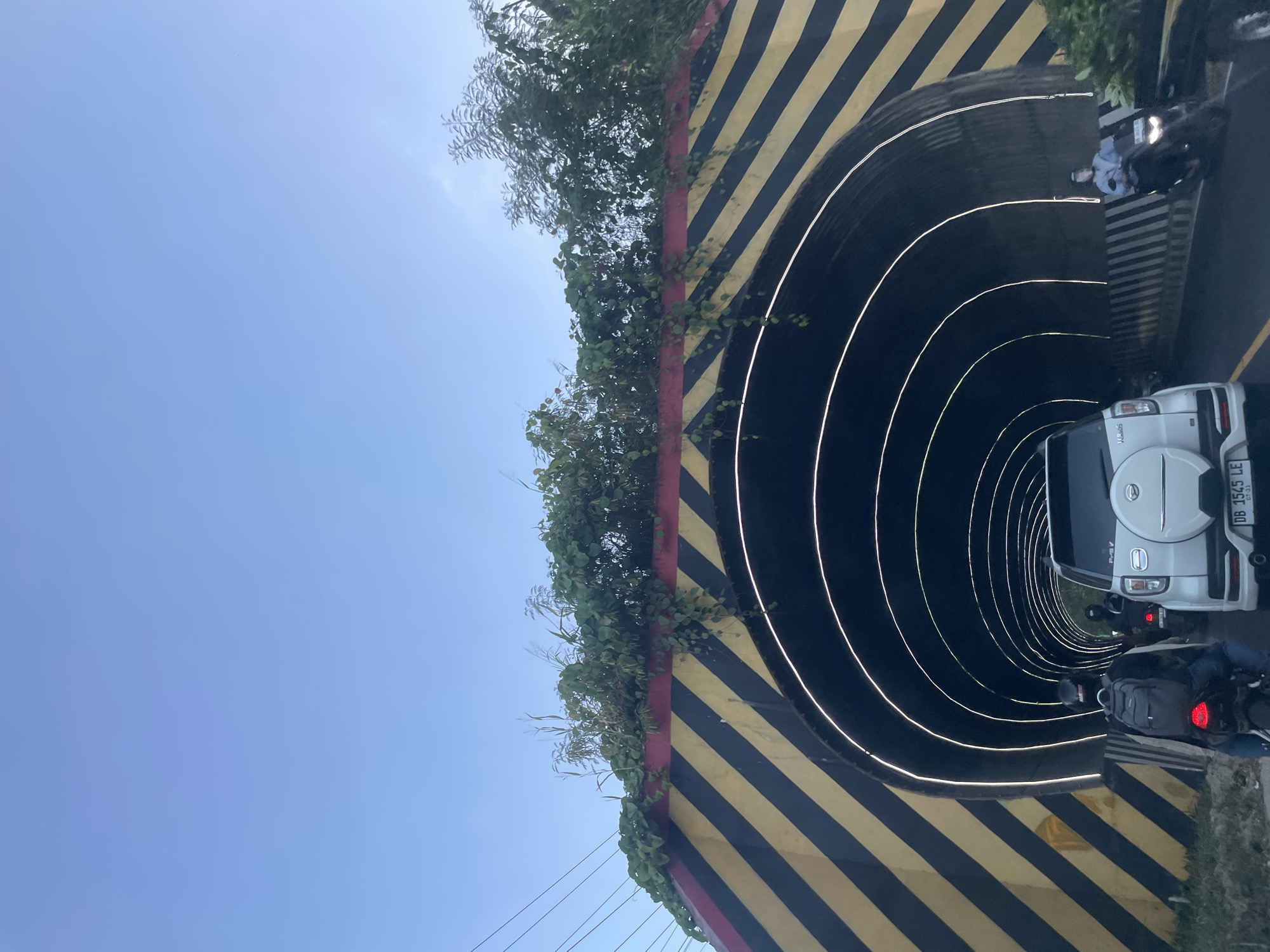 A white SUV is driving into a tunnel, with a motorcycle in front of it also entering the tunnel. The tunnel is illuminated by circular white lights along its ceiling. The exterior of the tunnel is painted with diagonal black and yellow stripes, with red trim at the top. Green vines and plants grow on top of the tunnel's structure. The sky above is clear and blue. The image appears to be taken from a vehicle driving on a road in Manado, Indonesia, likely near a section of road that passes through or under a structure.