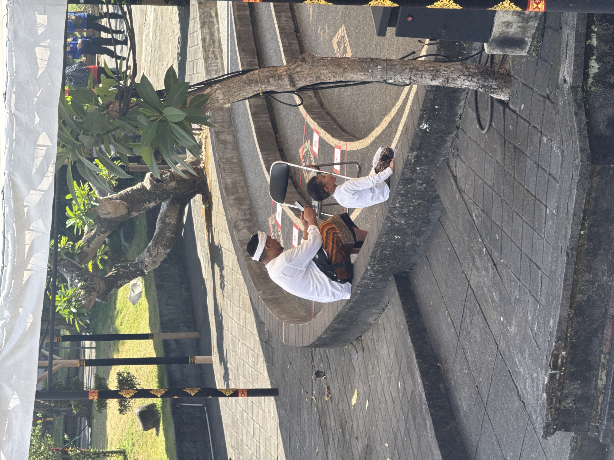 In the city of Denpasar, Indonesia, on a sun-drenched day, two individuals, an adult male and a young boy, are seated on the edge of a tiered, circular stone seating area. The adult, wearing a white long-sleeved shirt, a white head covering, and a patterned sarong, is looking down at a mobile phone in his hands. The young boy, dressed in a white long-sleeved shirt and patterned shorts, is looking forward, his legs crossed. A black folding chair is positioned behind the adult. The background features a well-maintained garden with grass, trees, and large stones. Several black poles decorated with gold patterns are visible, along with a white canopy overhead. The ground is paved with grey square tiles, and some fallen leaves are scattered on the surface.