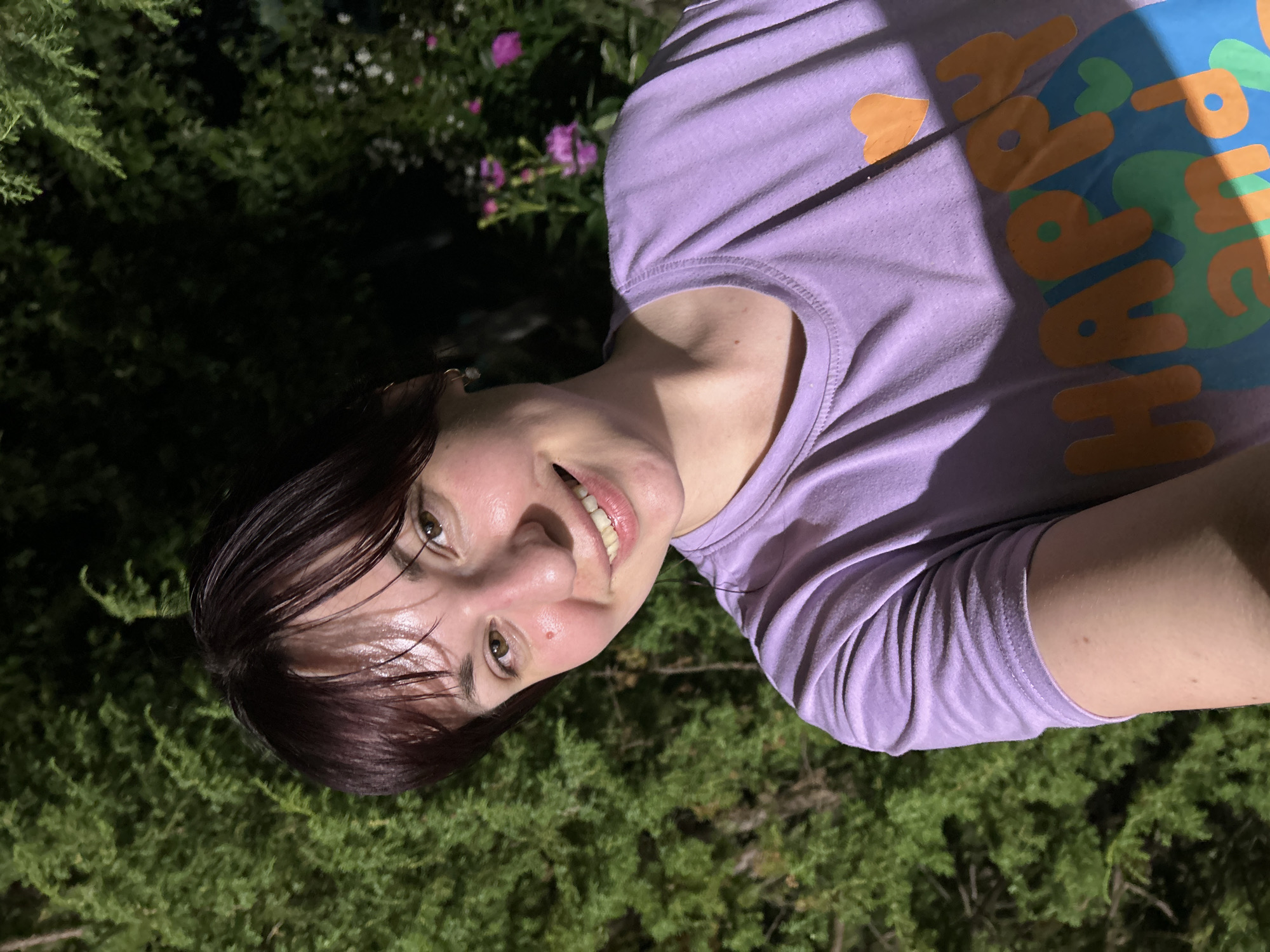 A woman with short, dark hair and a light complexion is smiling and looking directly at the camera. She is wearing a light purple t-shirt with an orange and blue graphic design that appears to say "Happy". The background is dark and out of focus, with visible green foliage, likely trees or bushes, and some pink flowers. The lighting suggests it is nighttime. The setting appears to be outdoors, possibly a garden or yard. The image quality is sufficient to discern facial features and clothing details. There is no indication of the specific location within Fokino, Russia, other than it is an outdoor environment.