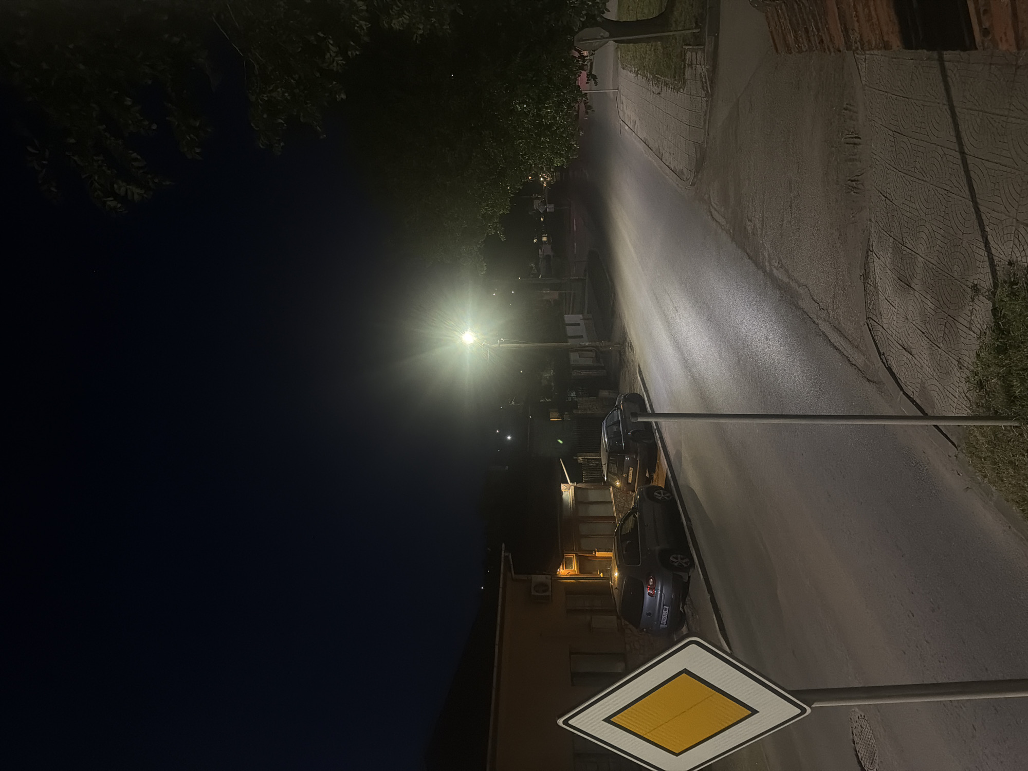 The image depicts a nighttime street scene, likely in a residential or mixed-use area in Iskar, Bulgaria. The dark sky indicates late evening or night. A prominent streetlight on the right side of the street emits a bright flare, illuminating the asphalt road surface which appears reflective. To the right of the road, a patterned sidewalk made of light-colored, textured pavers with circular and wavy designs is visible.
On the left side of the street, dark tree foliage is silhouetted against the night sky. Further down, several buildings are visible, with some windows illuminated, and an air conditioning unit can be seen on the exterior of one structure. Two cars are parked along the left side of the road. The closer vehicle is a dark-colored hatchback, and its license plate, "BA 6959 HM," is clearly legible.
In the immediate foreground, a yellow diamond-shaped "priority road" traffic sign with a white border is prominently displayed. No people are visible, and no specific activity is taking place, contributing to the quiet atmosphere of the scene.