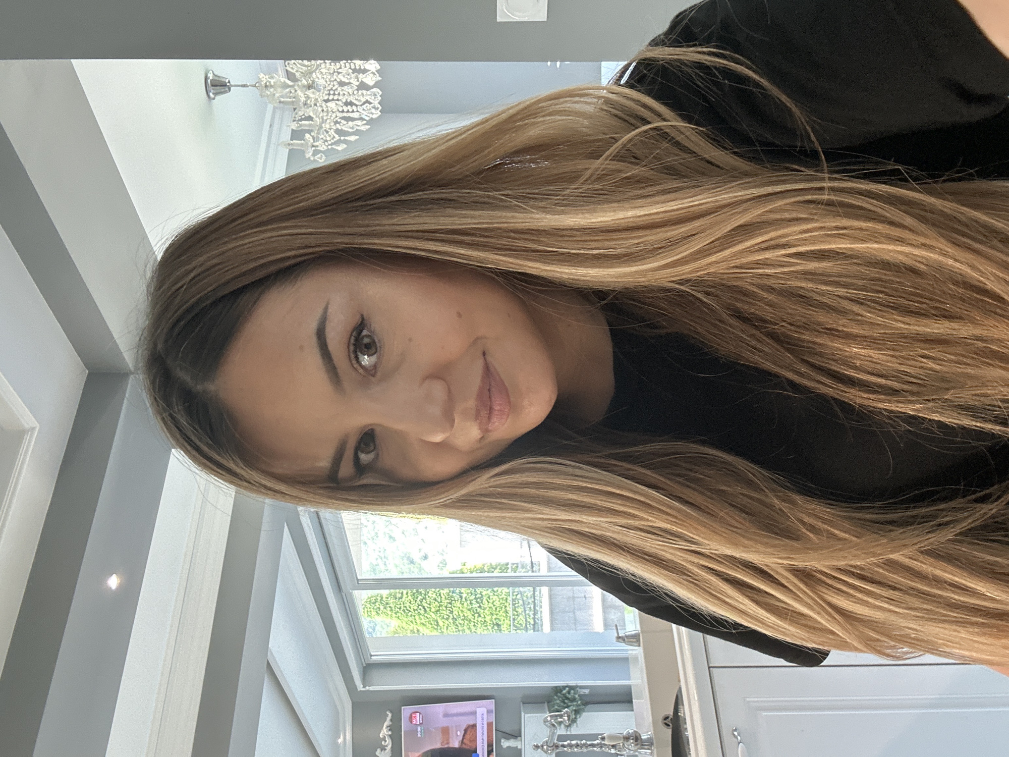 A young to middle-aged woman with long, layered, light brown hair with blonde highlights is depicted from the upper torso up, looking directly at the camera. Her hair is parted slightly to her left and falls over her shoulders and chest. She has brown eyes, well-defined eyebrows, and is wearing light makeup, including eyeliner. Her lips are softly closed in a subtle smile, showing a natural expression. She is wearing a black short-sleeved top.
The setting appears to be a contemporary interior space, likely a kitchen or living area, suggested to be in Asenovgrad, Bulgaria, given the discernible Bulgarian television channel and programming text visible on a flat-screen television in the background. To the left of the subject, white, possibly shaker-style, cabinetry is visible, along with a stainless steel sink and a decorative chrome faucet with white ceramic accents. Beyond this, a large sliding glass door or window unit opens to an exterior view of lush green foliage. On the far left, a television screen displays a program with Bulgarian text, including "BTV" and "20:00," indicative of local broadcasting. The ceiling features a multi-tiered design with white molding and gray recessed beams, one of which contains a spotlight. A prominent crystal chandelier hangs from the ceiling in the upper right portion of the frame. The walls are painted a light gray. The scene is well-lit, with ample natural light entering from the window, supplemented by interior artificial lighting.