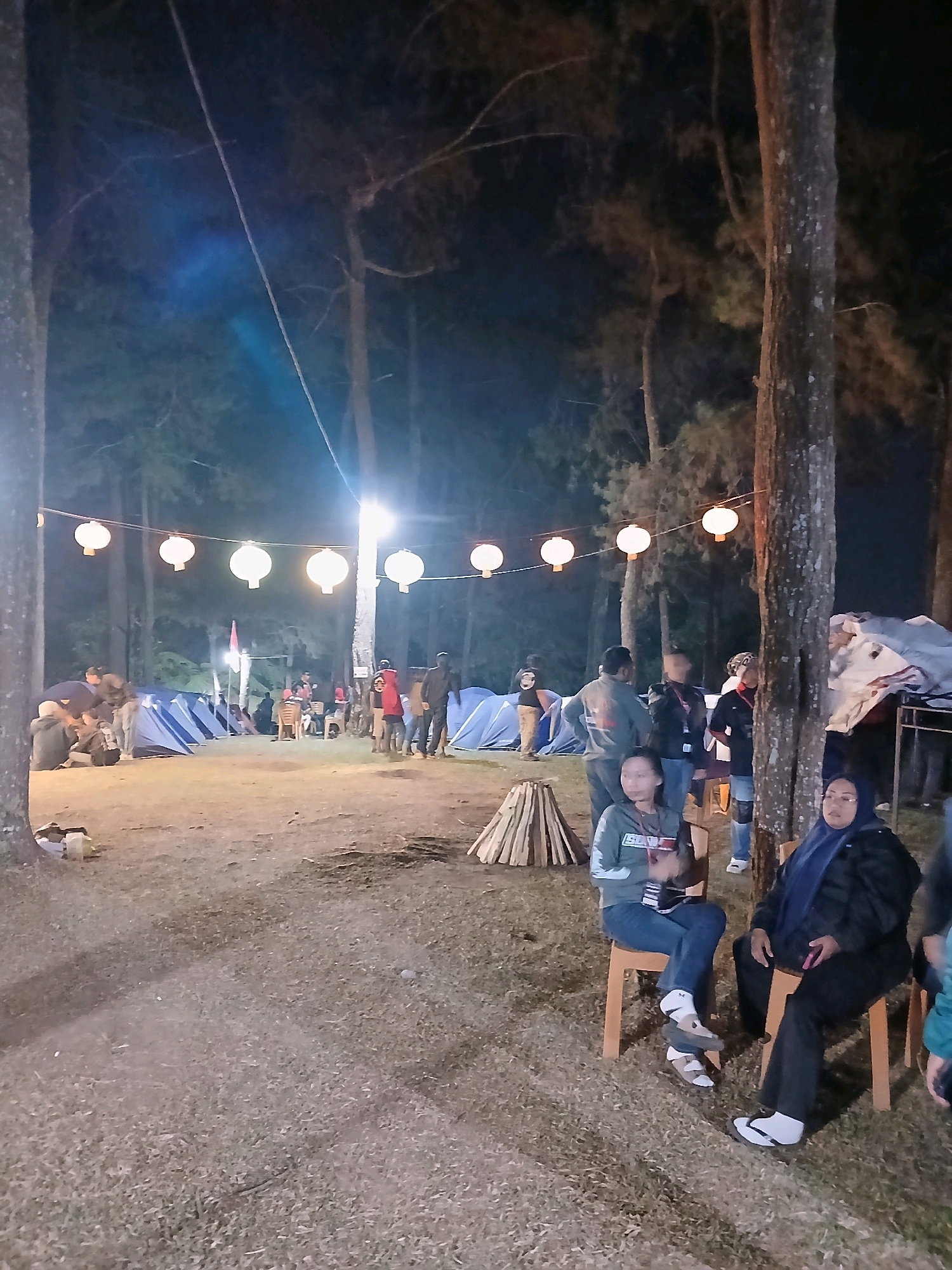 The image captures a nighttime scene at a campsite in Berjo, Indonesia, likely during a camping event or festival. Several tents are pitched on the ground, indicating a temporary settlement. Overhead, a string of white lanterns are illuminated, casting a warm glow on the surroundings. In the foreground, two women are seated on chairs. One woman, wearing a blue hijab and dark clothing, is looking towards the right. The other woman, wearing a grey sweatshirt and blue jeans, is looking down at her lap. A pile of firewood is situated to their left, suggesting preparations for a campfire. In the background, a group of people are gathered near the tents, some standing and some sitting. The environment appears to be a forested area, with tall trees framing the scene. The overall atmosphere suggests a communal gathering in an outdoor setting at night.