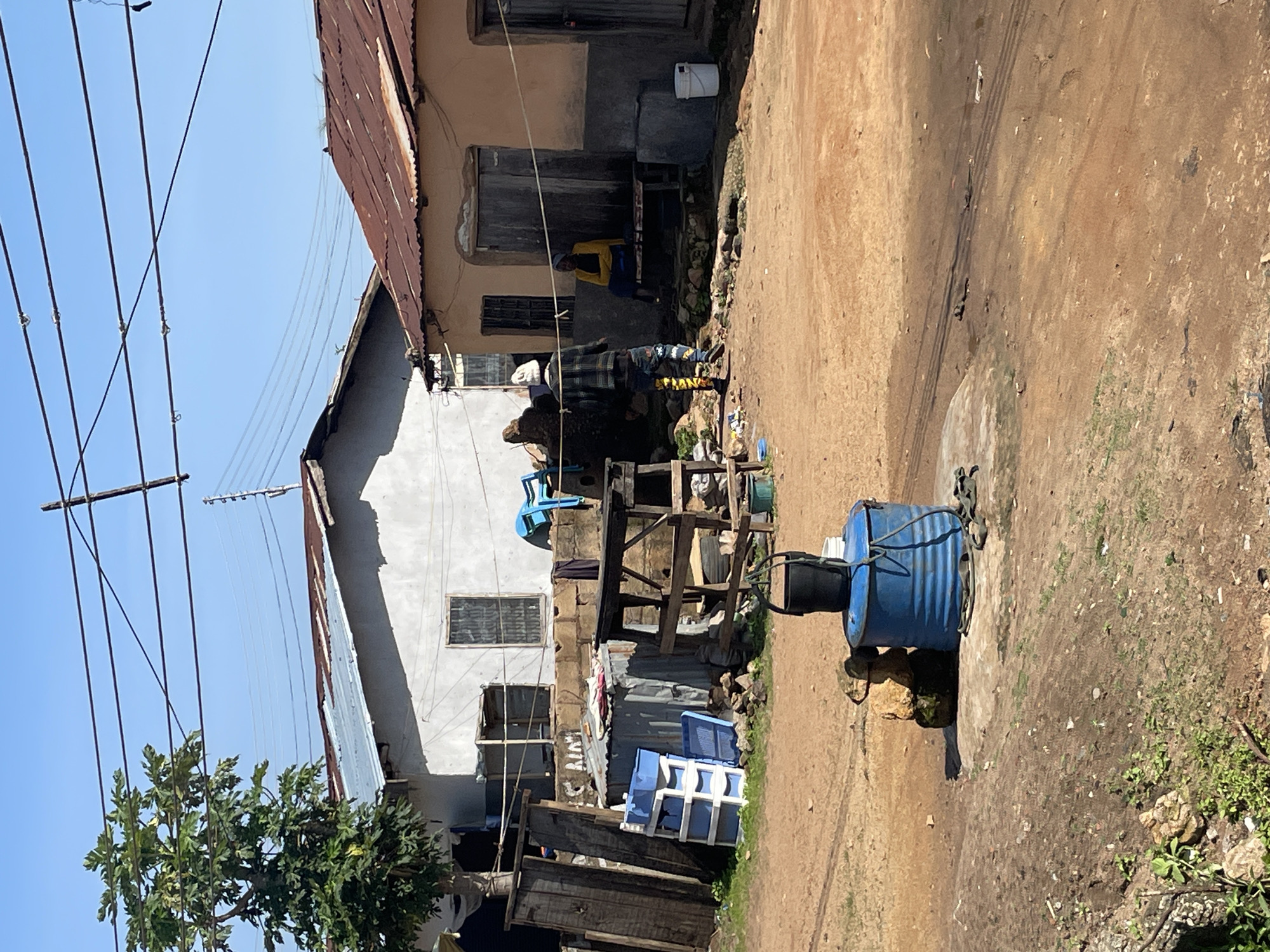 In Jos, Nigeria, an outdoor scene unfolds under a clear sky, with a dirt road dominating the foreground. To the left, a leafy green tree stands tall beside weathered wooden planks leaning against a gray, stucco building with a corrugated metal roof. In front of this building, a stack of blue plastic drawers sits next to a small corrugated metal shed.
Further into the scene, a rustic wooden table structure is visible. On the right side of the frame, a peach-colored building with a rusty corrugated metal roof features a wooden door and a barred window. A woman in a yellow shirt and blue skirt sits on a bench outside this building.
A person wearing a patterned jacket and vibrantly patterned pants stands facing away from the viewer, looking towards the seated woman. In the center of the image, a blue barrel holds a black bucket, supported by stacked rocks. The ground is a mix of dry dirt and sparse green vegetation. Overhead, electrical wires crisscross the sky.
