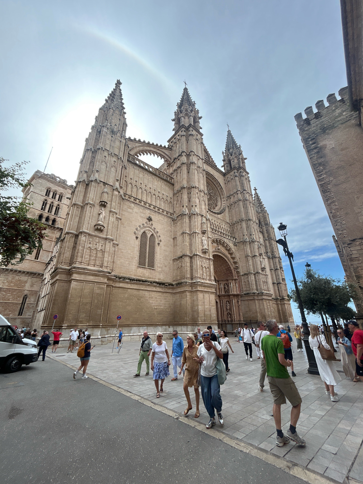 The image shows a wide-angle, eye-level view of the Palma Cathedral (La Seu) in Palma de Mallorca, Spain, on a sunny day. The cathedral, made of sandstone, dominates the background with its Gothic architecture, featuring spires, arched windows, and a large rose window. A faint rainbow is visible in the sky above the cathedral. In the foreground and midground, a bustling pedestrian area lines the street in front of the cathedral. Numerous tourists, dressed in casual summer clothing, are walking, standing, and interacting. Some are looking at the cathedral, others are in conversation. A white van is parked on the left side of the street. Streetlights and trees line the pedestrian walkway. The ground is paved with stone tiles. The overall scene depicts a popular tourist attraction with a lively atmosphere.