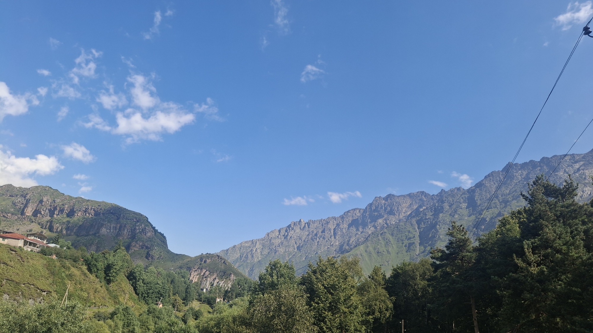 The image depicts a daytime outdoor scene featuring a mountain range under a bright blue sky with scattered white clouds. The foreground is dominated by lush green trees, with some appearing to be deciduous and others coniferous. On the left side of the image, the slopes of a mountain are visible, with several buildings nestled on them, suggesting a residential area. These buildings have red tiled roofs and appear to be traditional houses. The middle ground and background are filled with large, rugged mountains, characterized by rocky outcrops and green vegetation on their lower slopes. The texture of the mountain faces suggests geological formations. In the upper right corner, dark wires, possibly power lines, stretch across the frame, indicating human infrastructure in the natural landscape. The overall environment suggests a rural or mountainous region.
