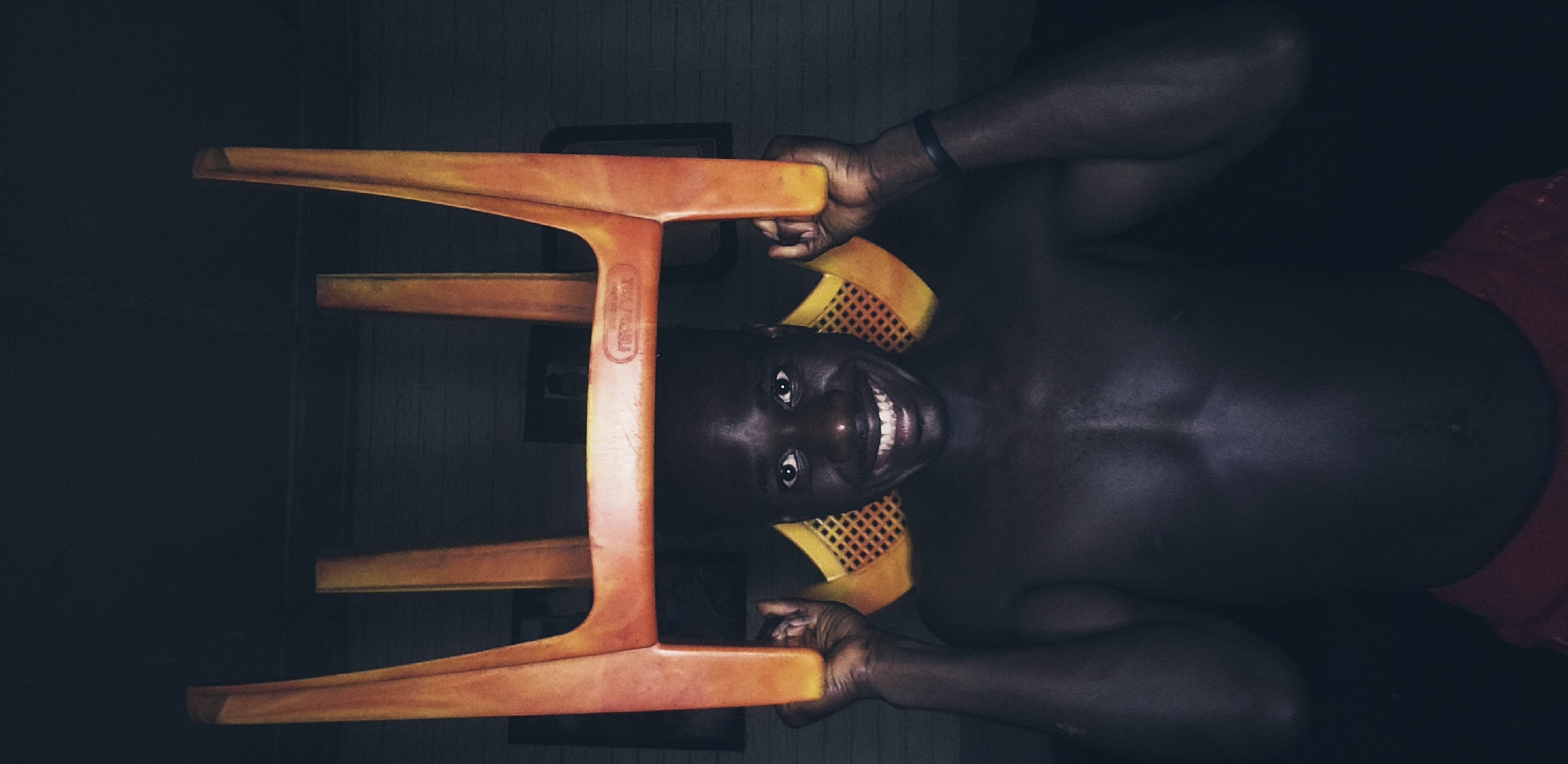 A shirtless dark-skinned man with a wide smile and white teeth holds an orange plastic chair over his head. The chair is positioned horizontally, with its legs pointing upwards and outwards. The man's hands are gripping the sides of the chair, with his thumbs on top and fingers underneath. He is looking directly at the camera. His torso and upper body are visible, and he appears to be wearing red shorts that are not fully in view. The background is dark and appears to be an indoor setting, with a brick wall texture visible behind the man and some framed pictures on the wall. The lighting is dim, with the man's face and upper body illuminated. The image was taken in Narayi, Nigeria.