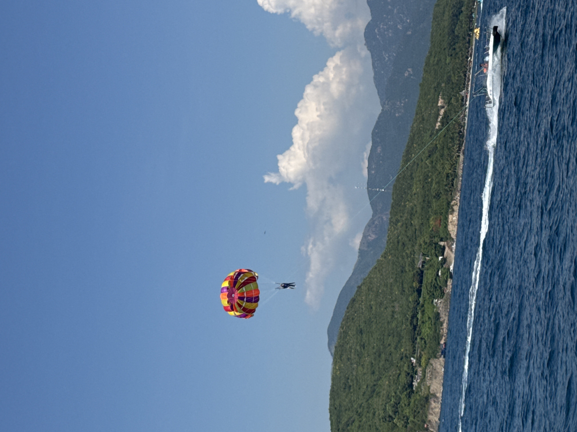 A parasailer with a colorful canopy is suspended in the air against a clear blue sky, with a chain of mountains covered in lush green vegetation forming the backdrop. Below the parasailer, a speed boat is cutting through the deep blue water, leaving a white wake. The boat appears to be towing the parasailer. The scene suggests a tourist activity likely in a coastal area. The presence of the mountains and the boat indicates a likely location in Bac Nha Trang Ward, Vietnam, a coastal city known for its beaches and water sports. The weather appears to be sunny and calm, suitable for outdoor activities.
